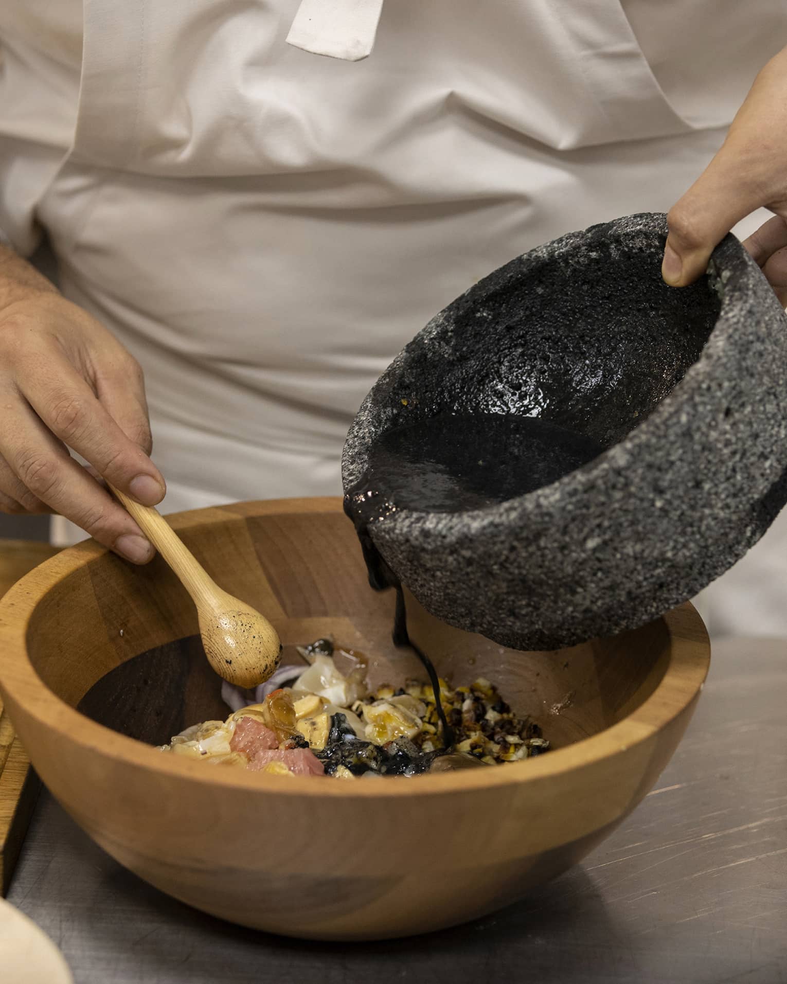 Chef pours black sauce from a stone bowl into a wood bowl of ingredients, a small wooden spoon in the other hand. 