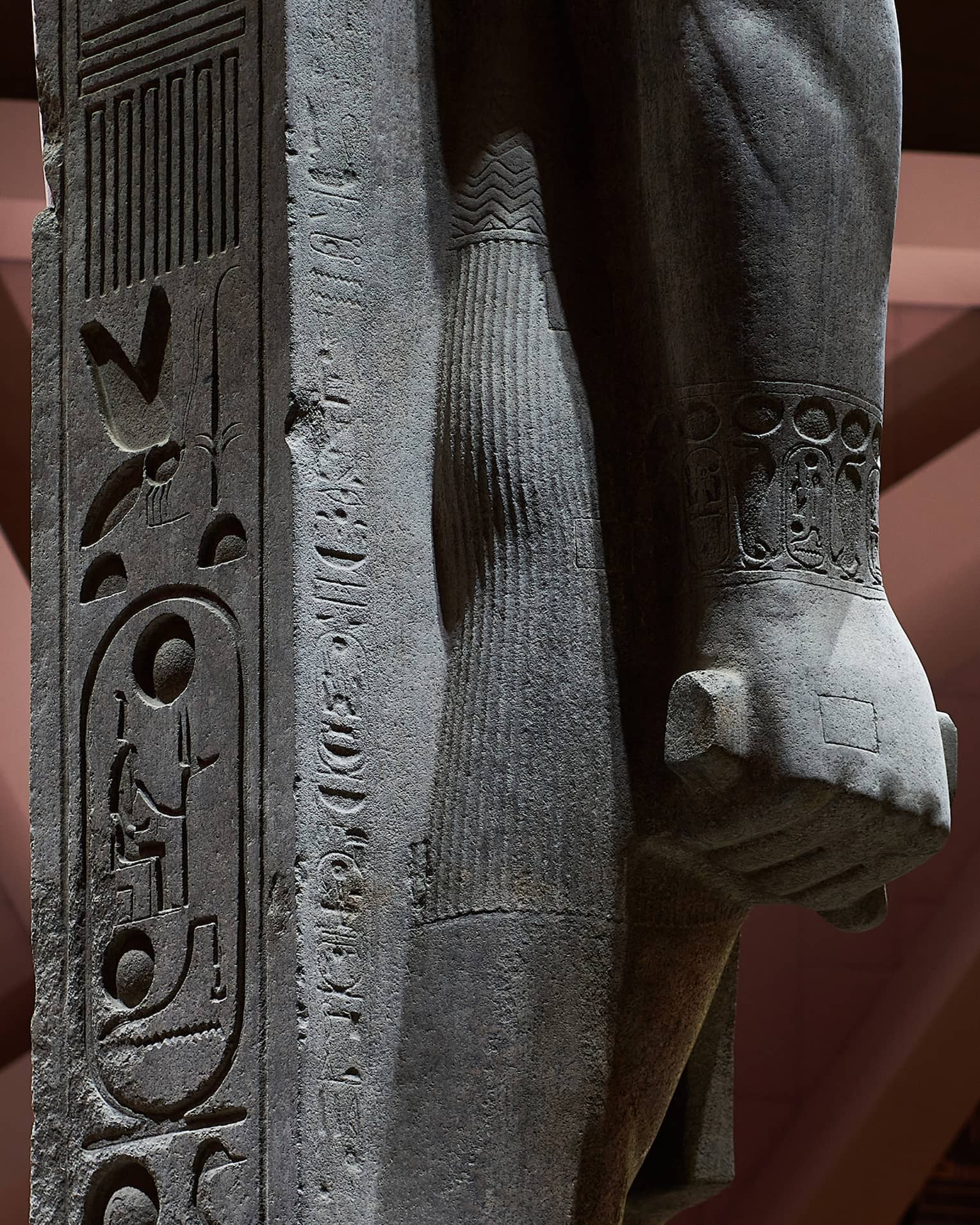 Close-up view of a statue's hand in a fist, supported by a stone inscribed with hyroglyph carvings. 