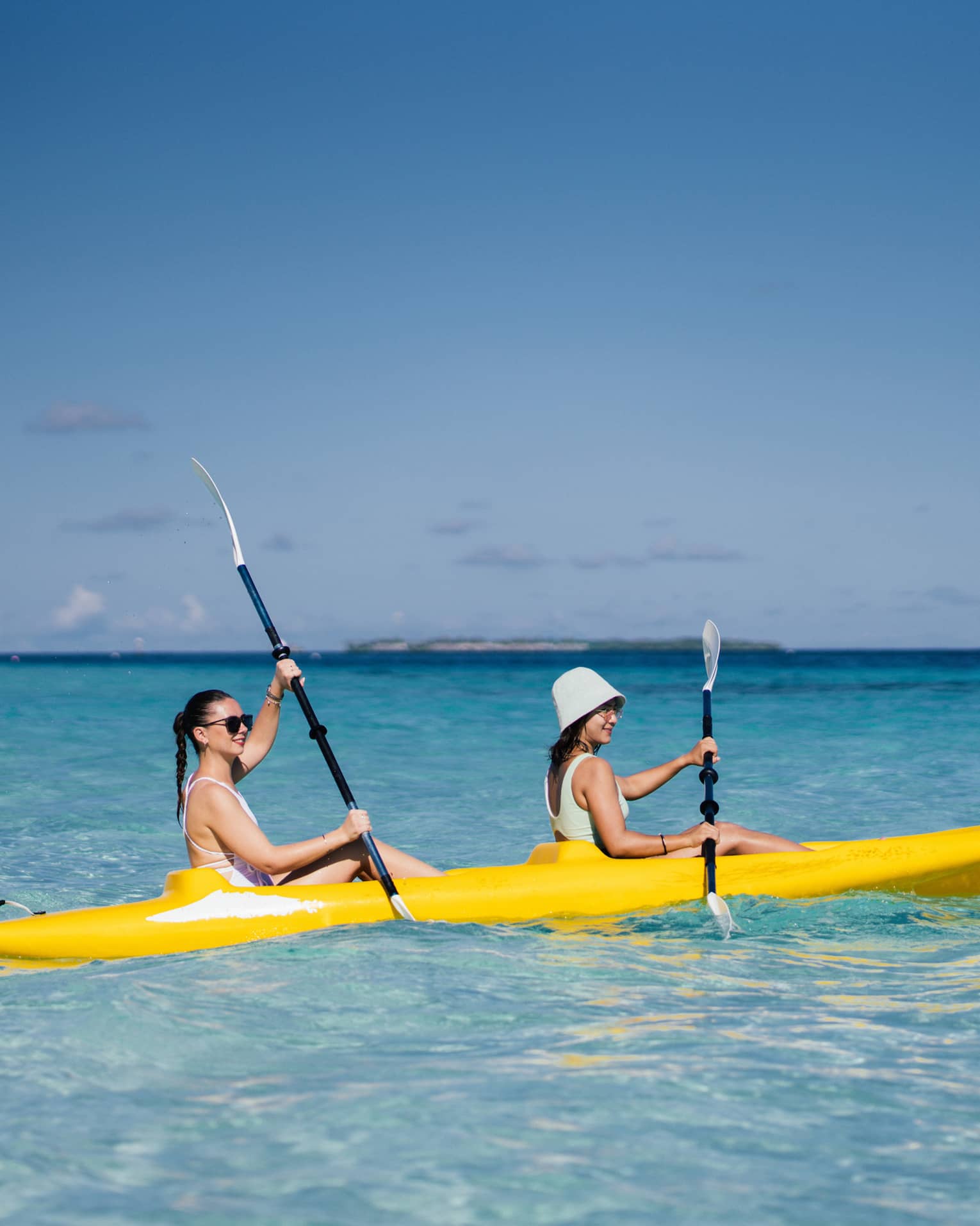Two people smile as they paddle a yellow kayak in a turquoise lagoon under a clear blue sky; low island in the distance.