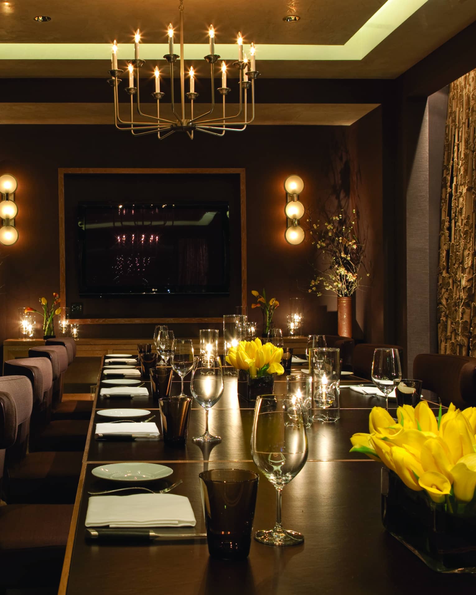 Long private dining table with plush chairs set with wine glasses, yellow flowers, candles, low lights on walls