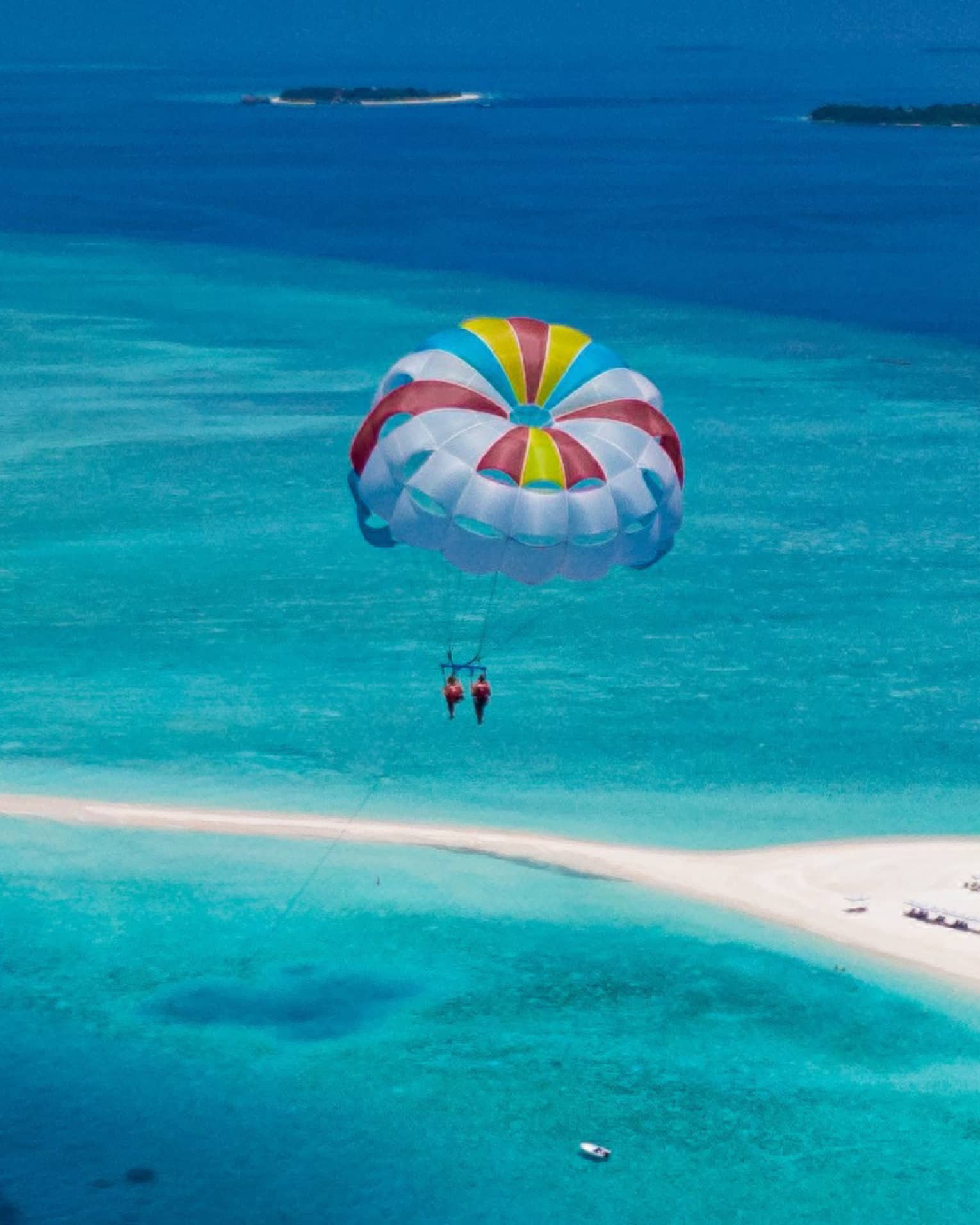 Aerial view of two people parasailing in tandem, colourful sail floating above an expanse of aqua water and a white sandbar.