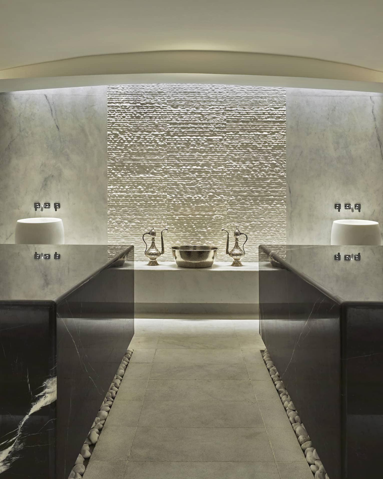 The interior of a hammam with grey marble walls, two black marble tables and a gold sink in the backfround