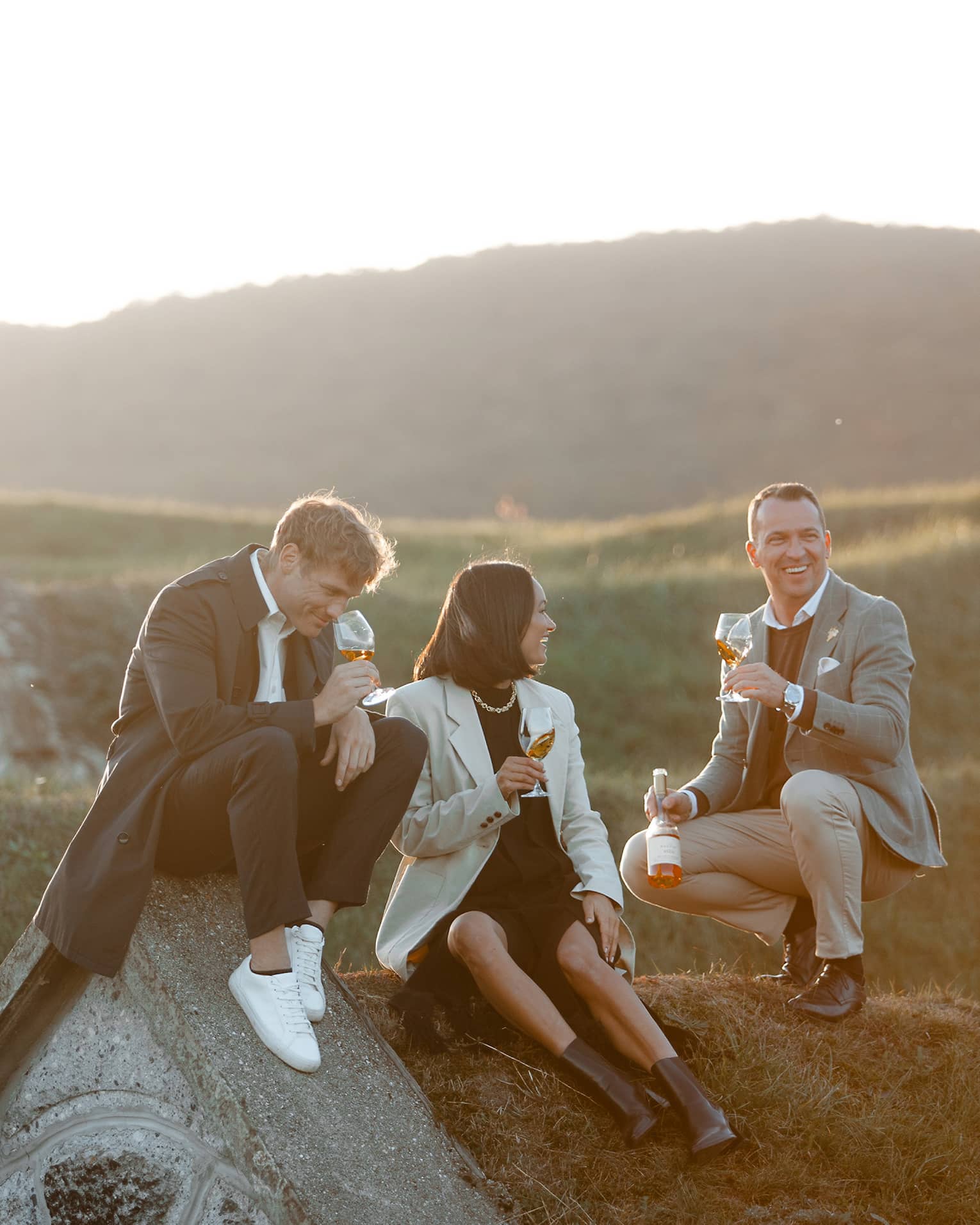 Three friends sitting on a hillside, smiling and drinking wine