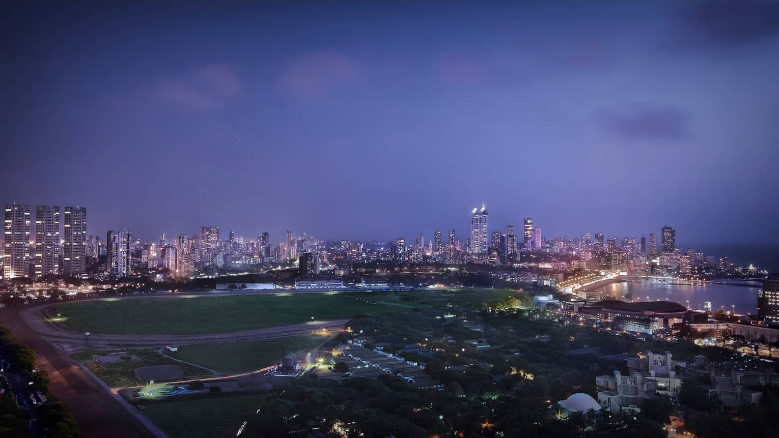 View over large field, track, Mumbai city skyline with buildings, lights at dusk