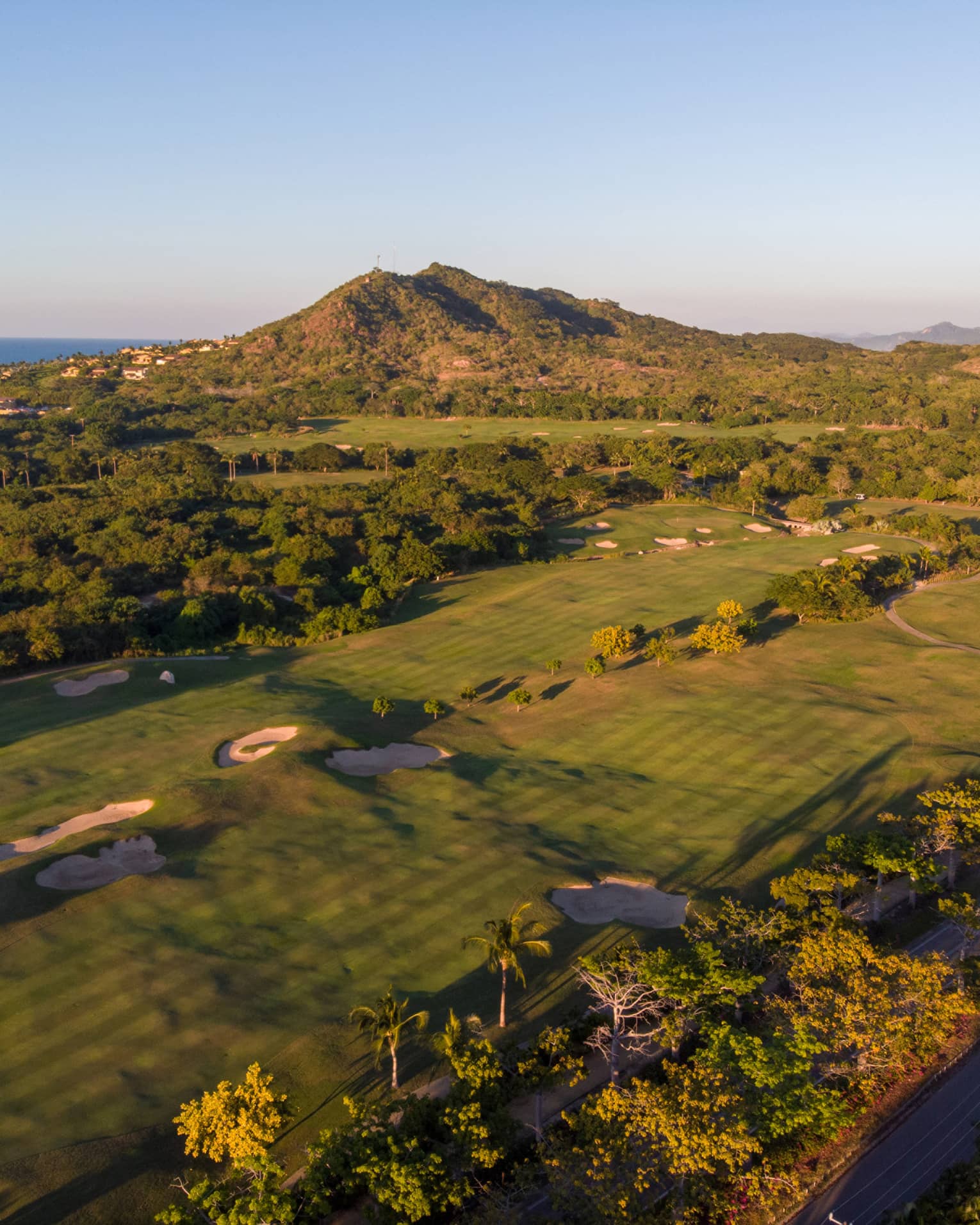 Aerial view of a golf course over the rolling coastal landscape; houses rising along the hillside, ocean in the distance.