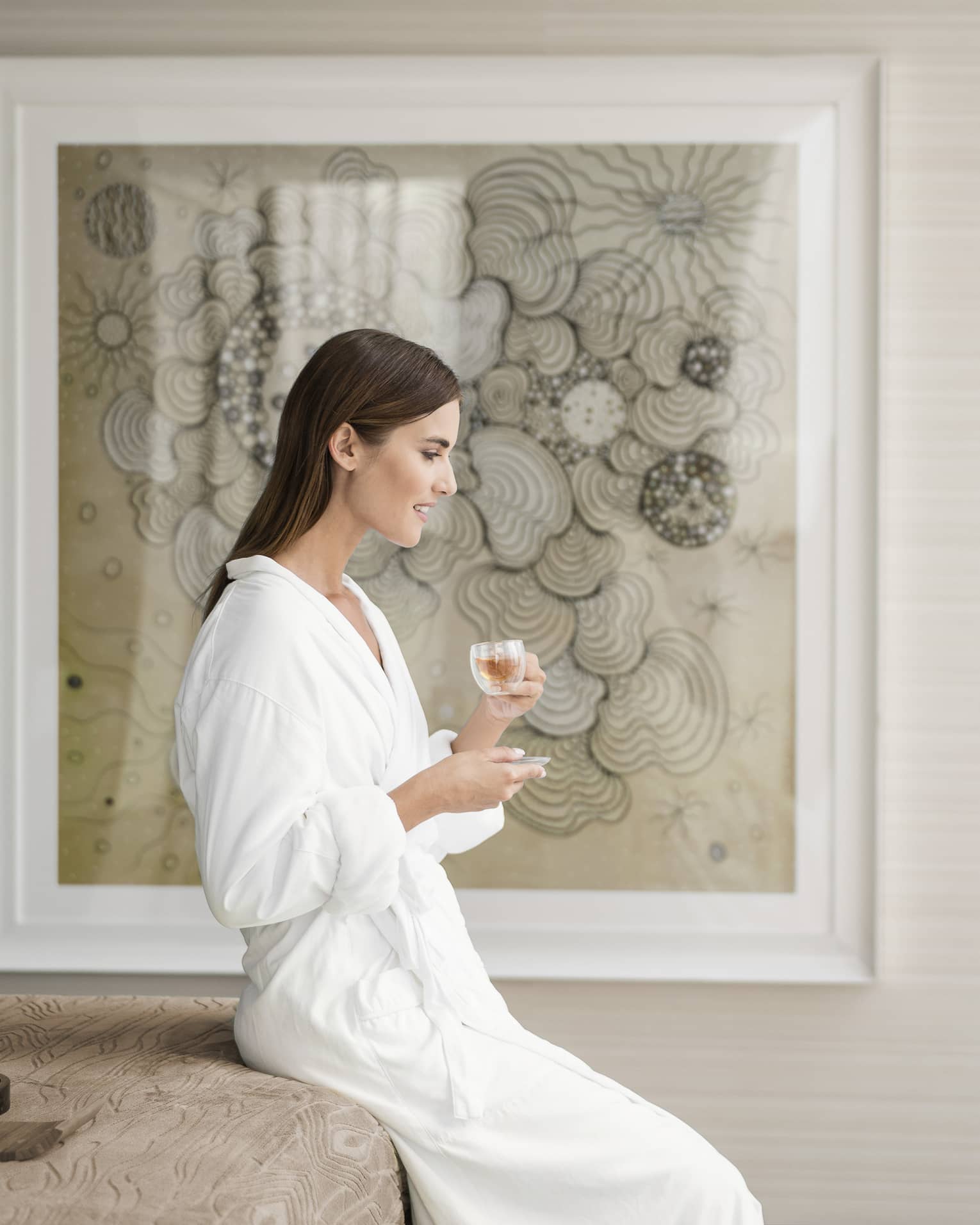 A woman in a white robe sipping from a small glass in front of a framed portrait.