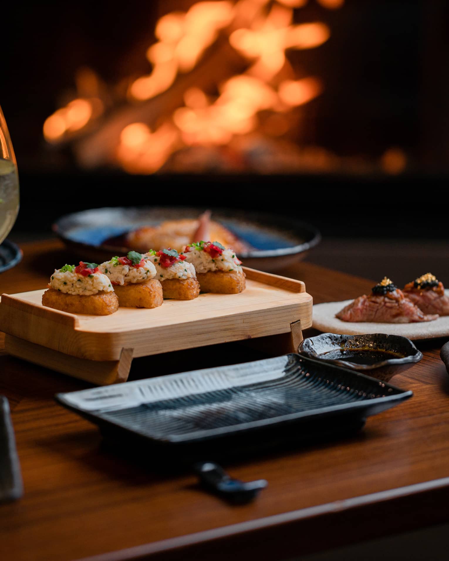 Sushi dinner setup on table with glass of white wine and sparkling fire in backdrop