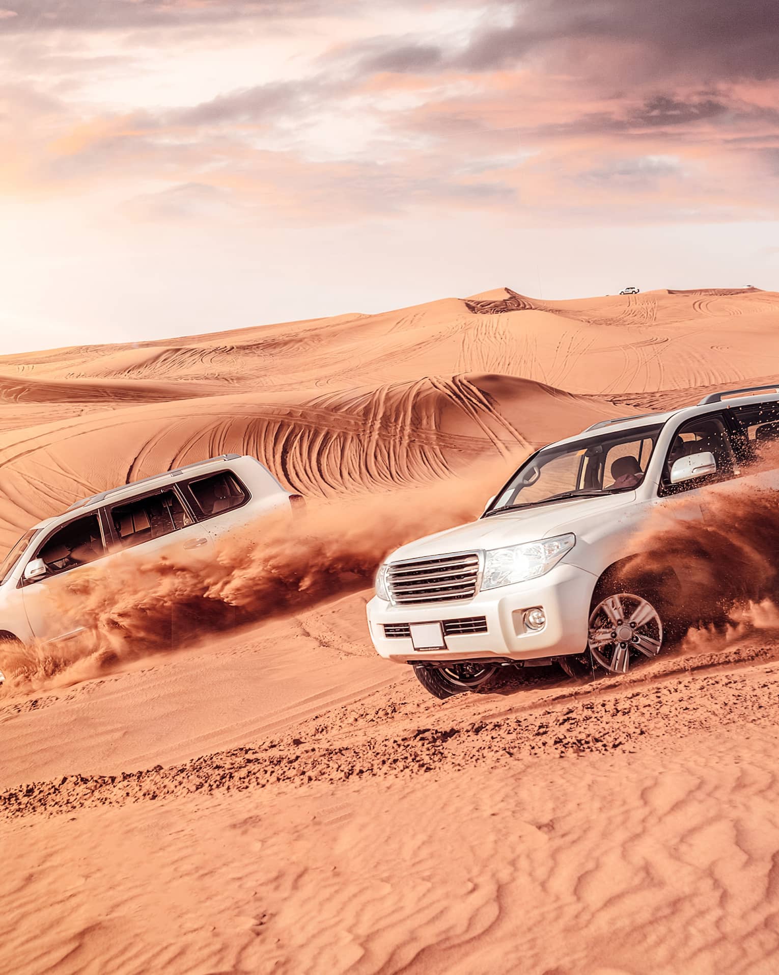 Sand flies from tires of two white SUV's driving across dunes