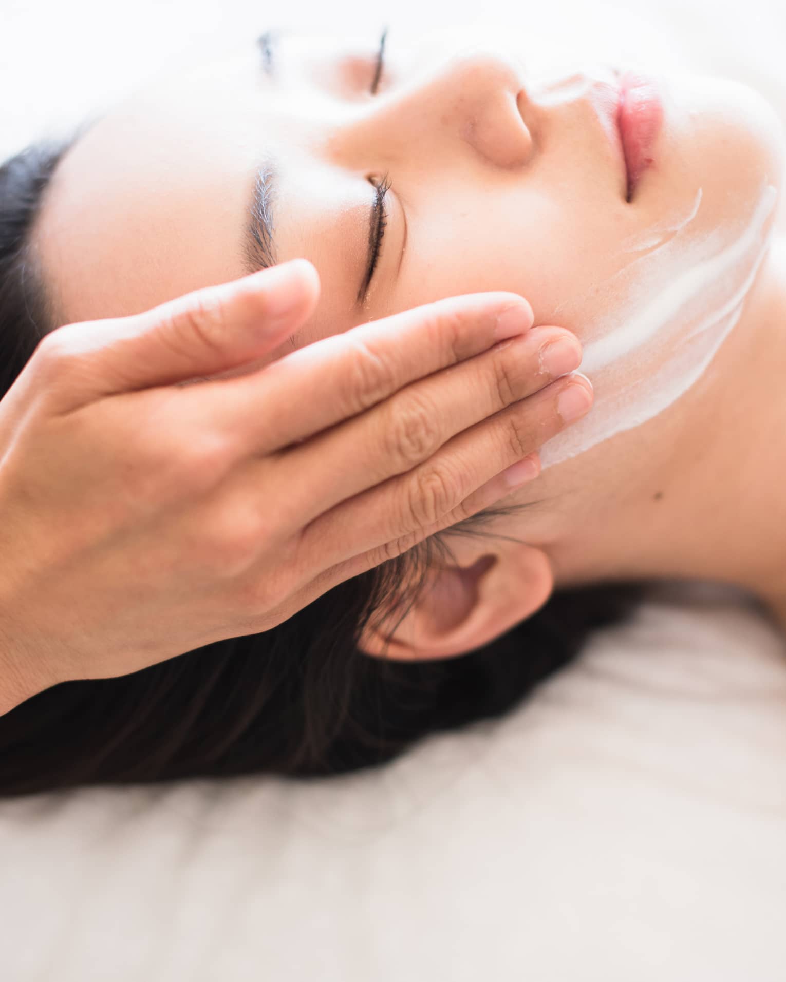 Close-up of spa attendant's hands rubbing white cream on woman's face as she lies on massage table