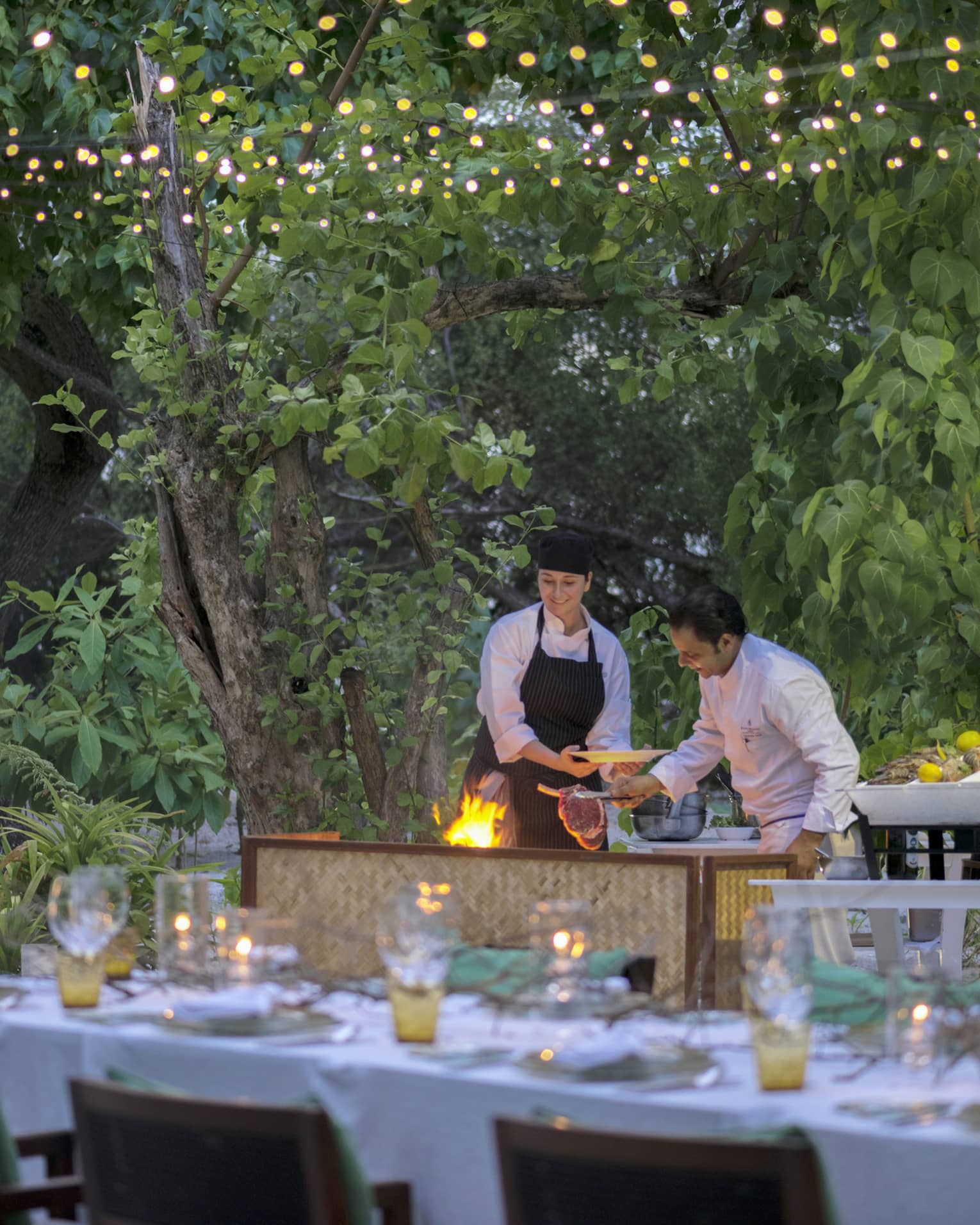 Chefs put large steak on flaming grill by long outdoor dining table, patio lights at night