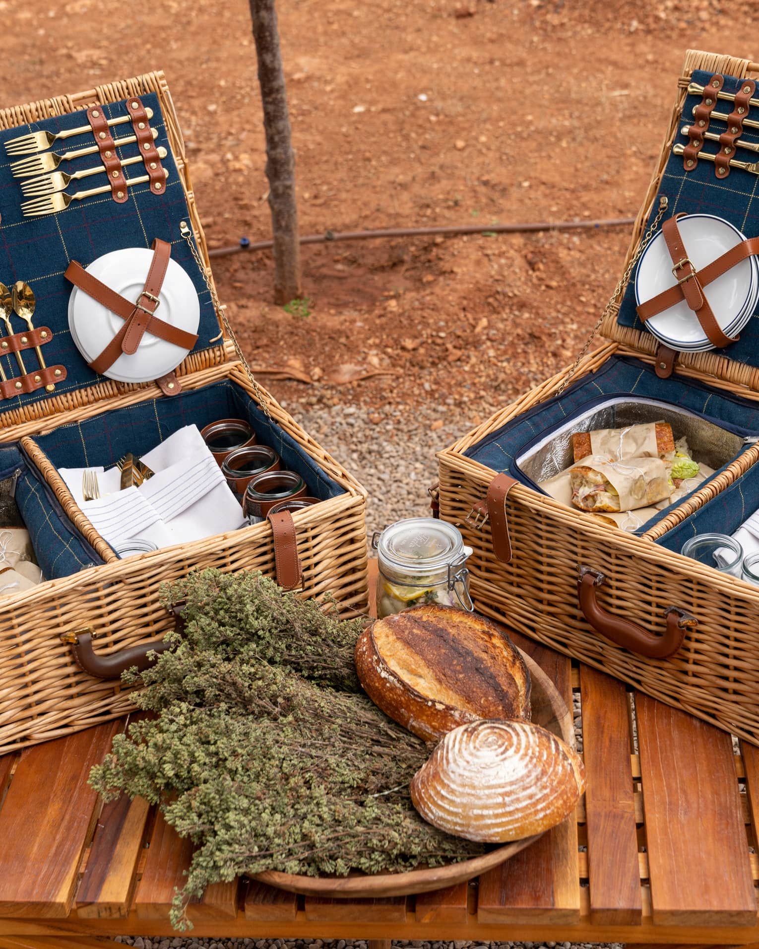 Plates and gold cutlery secured to the lids of picnic baskets containing parchment-wrapped sandwiches, napkins, and glasses.