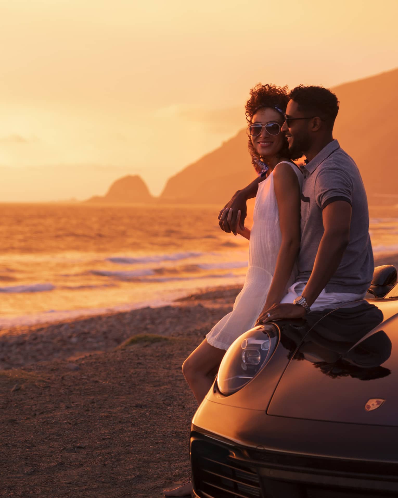 A couple leaning on a sports car that is parked on the beach with the ocean beside them.