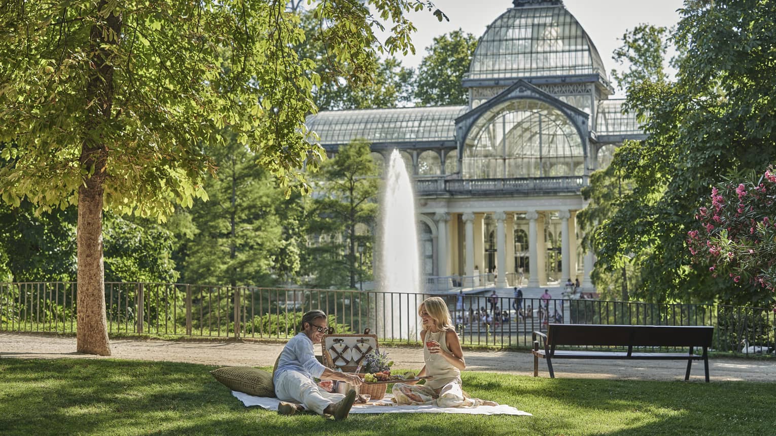 Couple on blanket on grass in Retiro Park, having picnic by park bench and near fountain