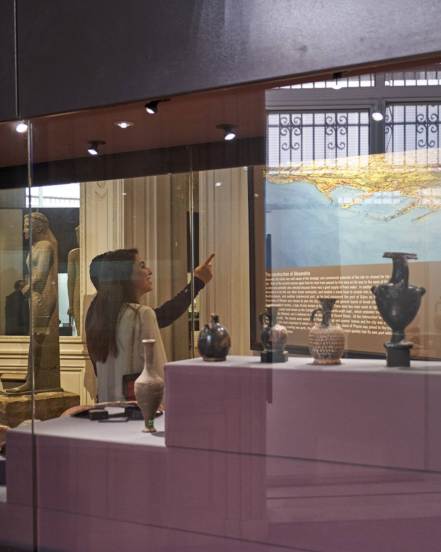 A visitor in a museum points up at a historical map lit up on a wall, behind them a display case with various vases and jugs.