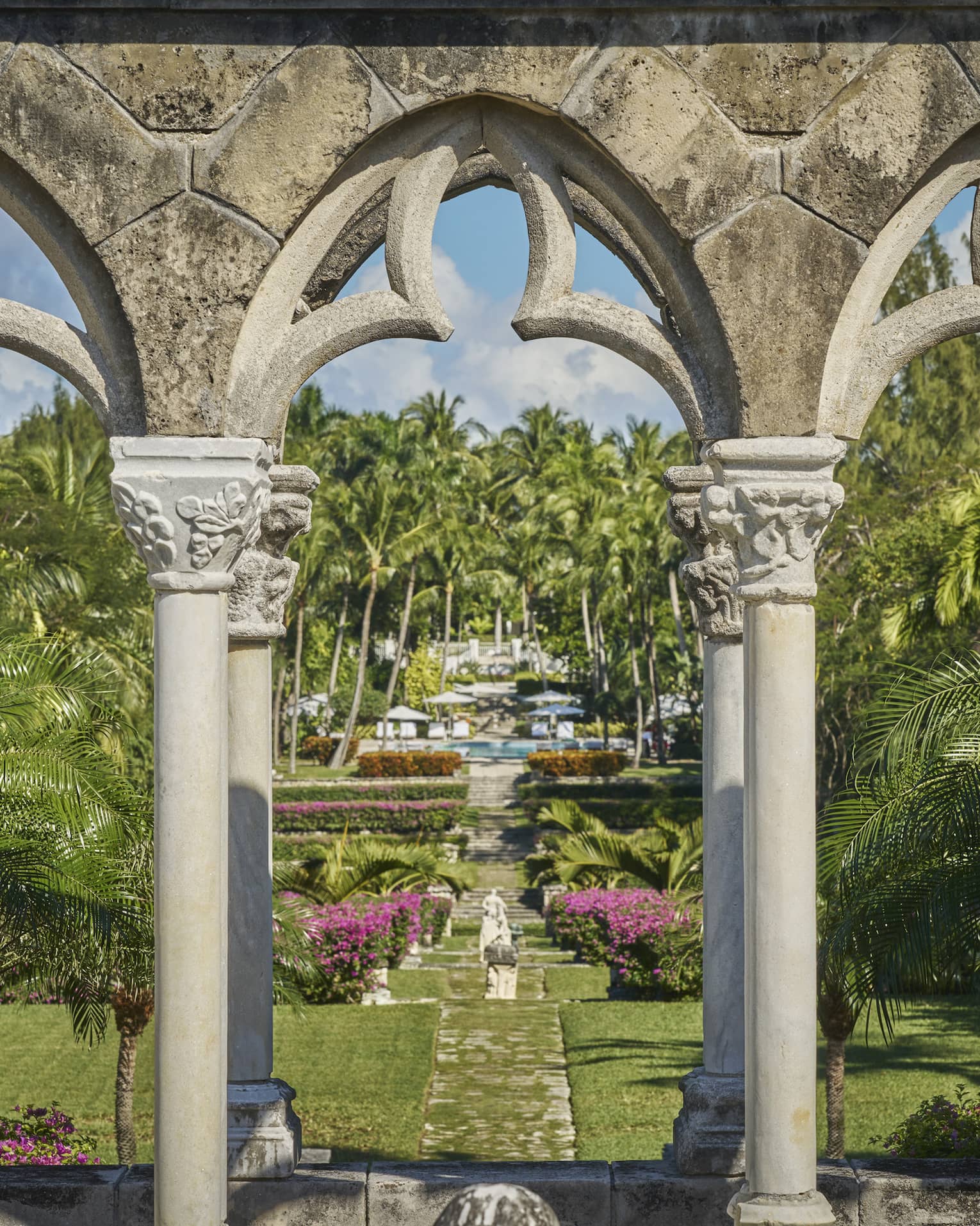 Stone arches leading to a green garden with grass, flowers and palm trees.