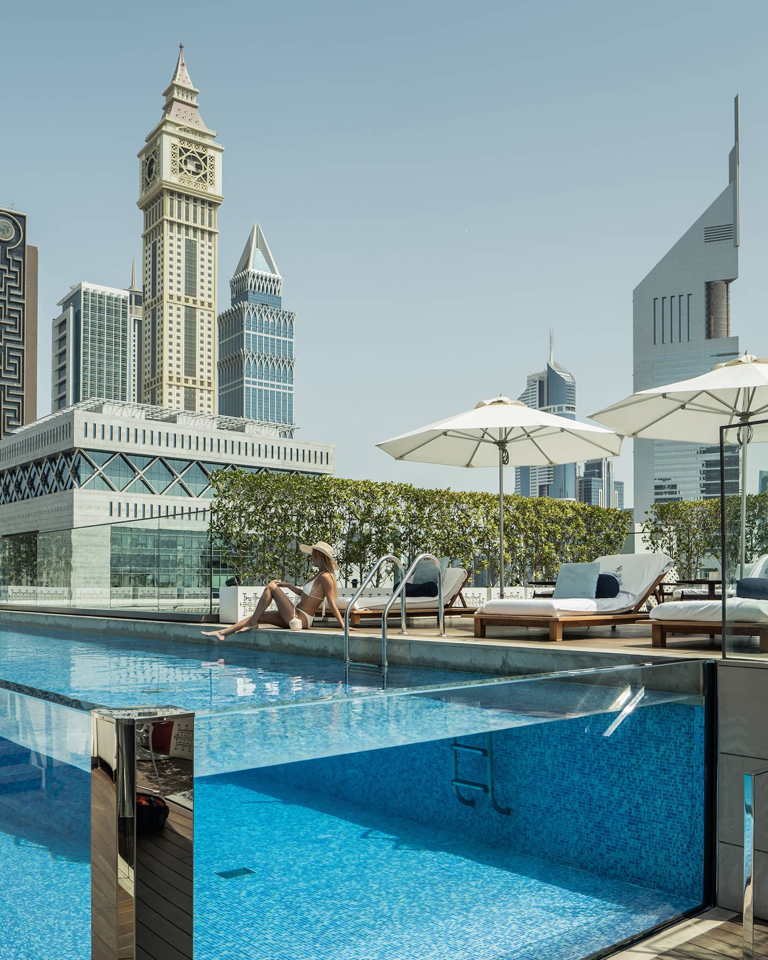 A guest lounges by a transparent rectangular rooftop pool, modern towers with geometric designs rising into the azure sky.