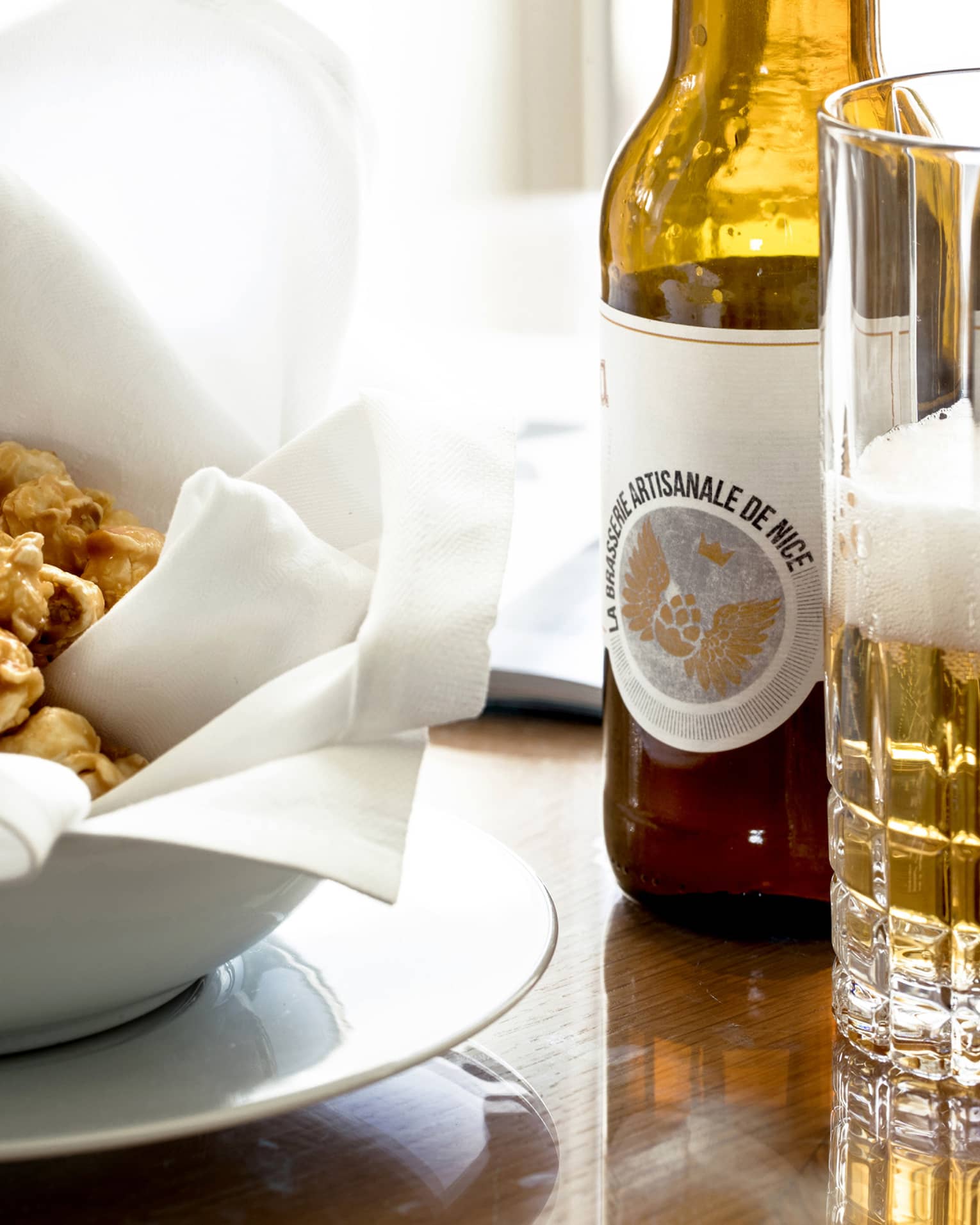 An amber bottle of artisanal beer beside a partially filled, foamy glass and a bowl of caramel popcorn on a cloth napkin. 