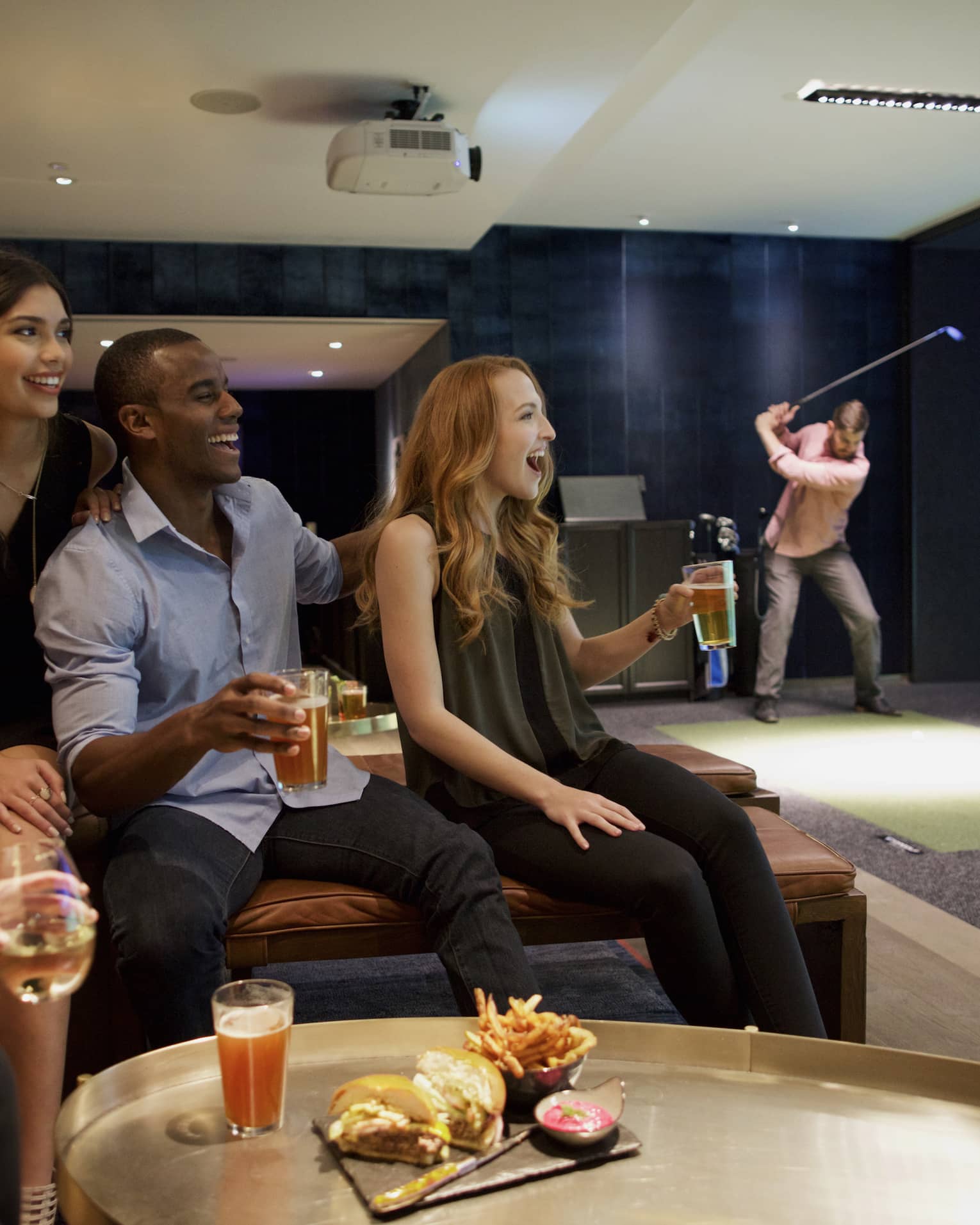 A man and three woman sip beer and cheer on a sports team in the foreground, while another man golfs in the background of a function room at four seasons hotel houston 