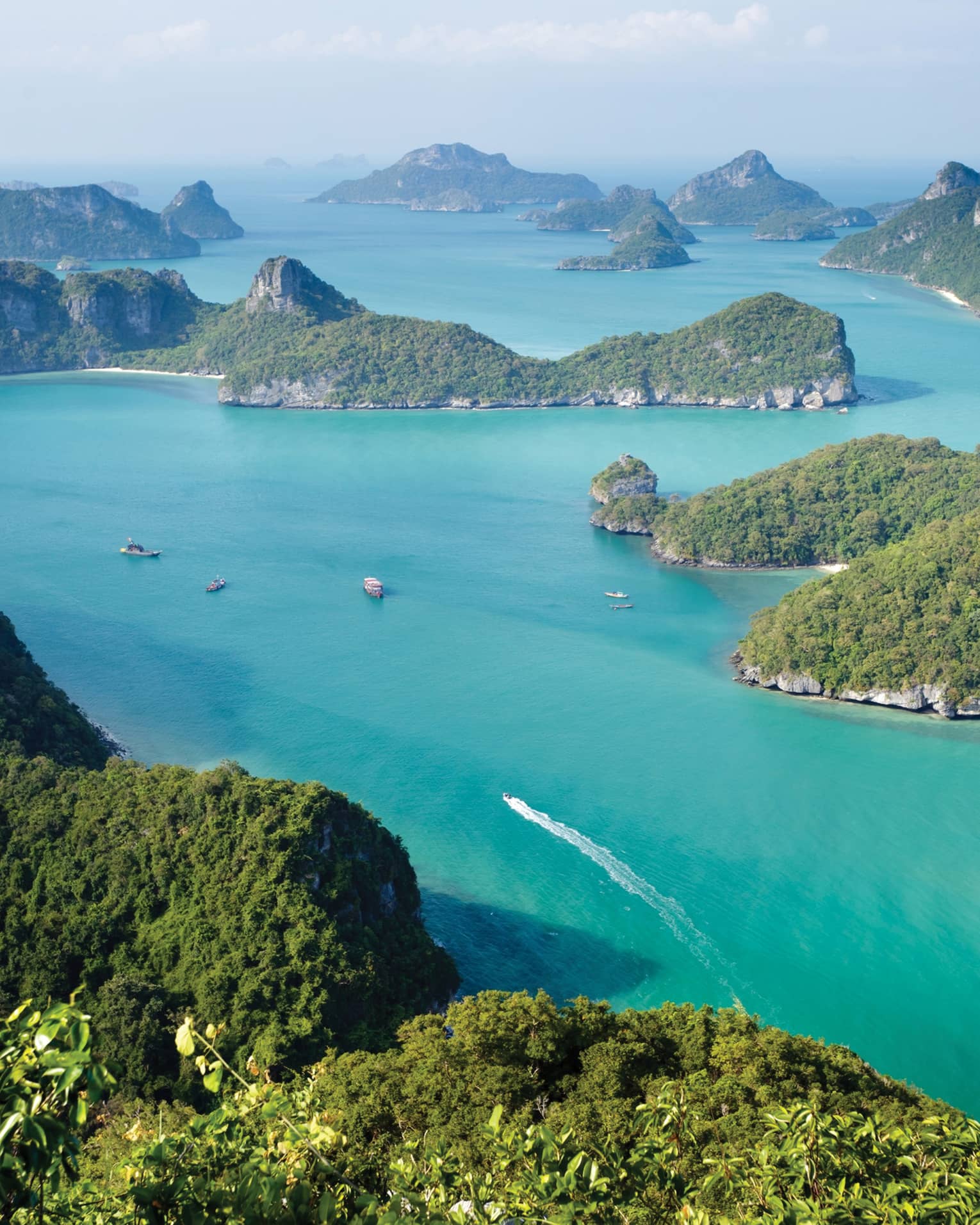 Aerial view of a dozen lush, forested, hilly islands of different sizes surrounded by calm turquoise water dotted with boats.