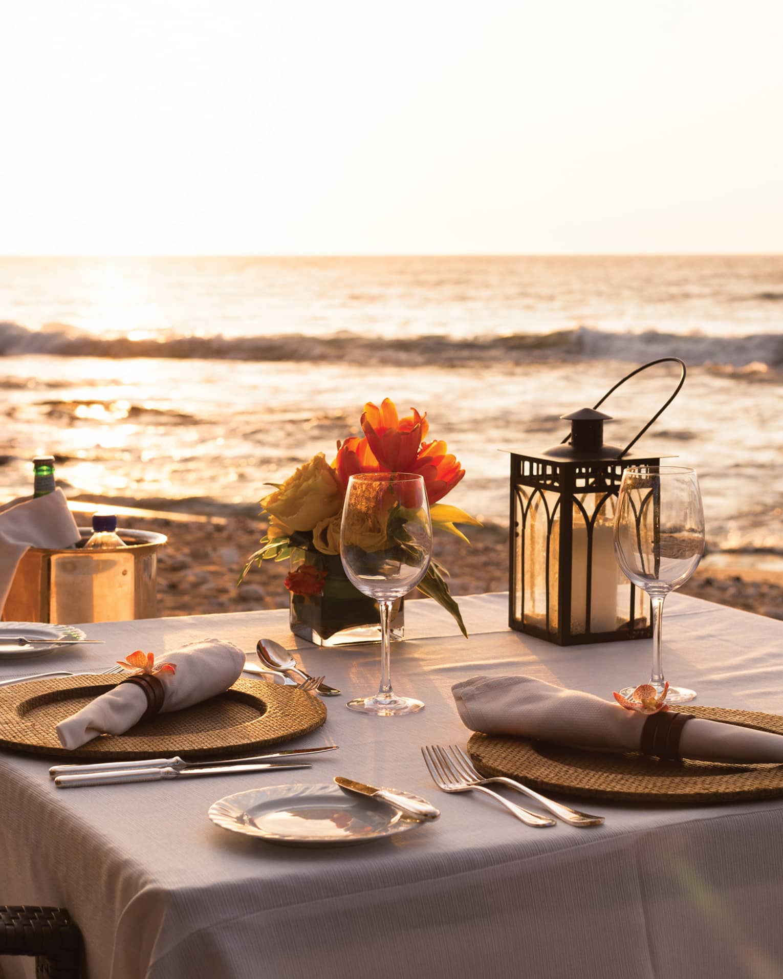 Close-up of formal place setting on table with white cloth, Champagne in bucket on beach at sunset