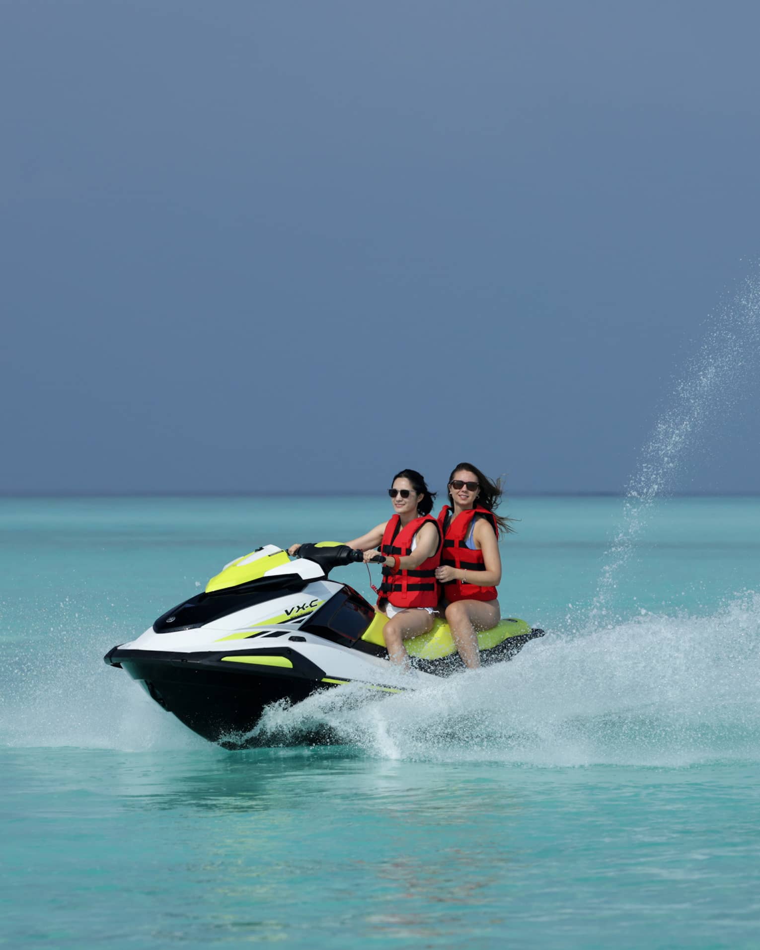 Wearing life jackets, two smiling jet-skiers ride through the clear ocean, water spurting up from beside and behind the boat.