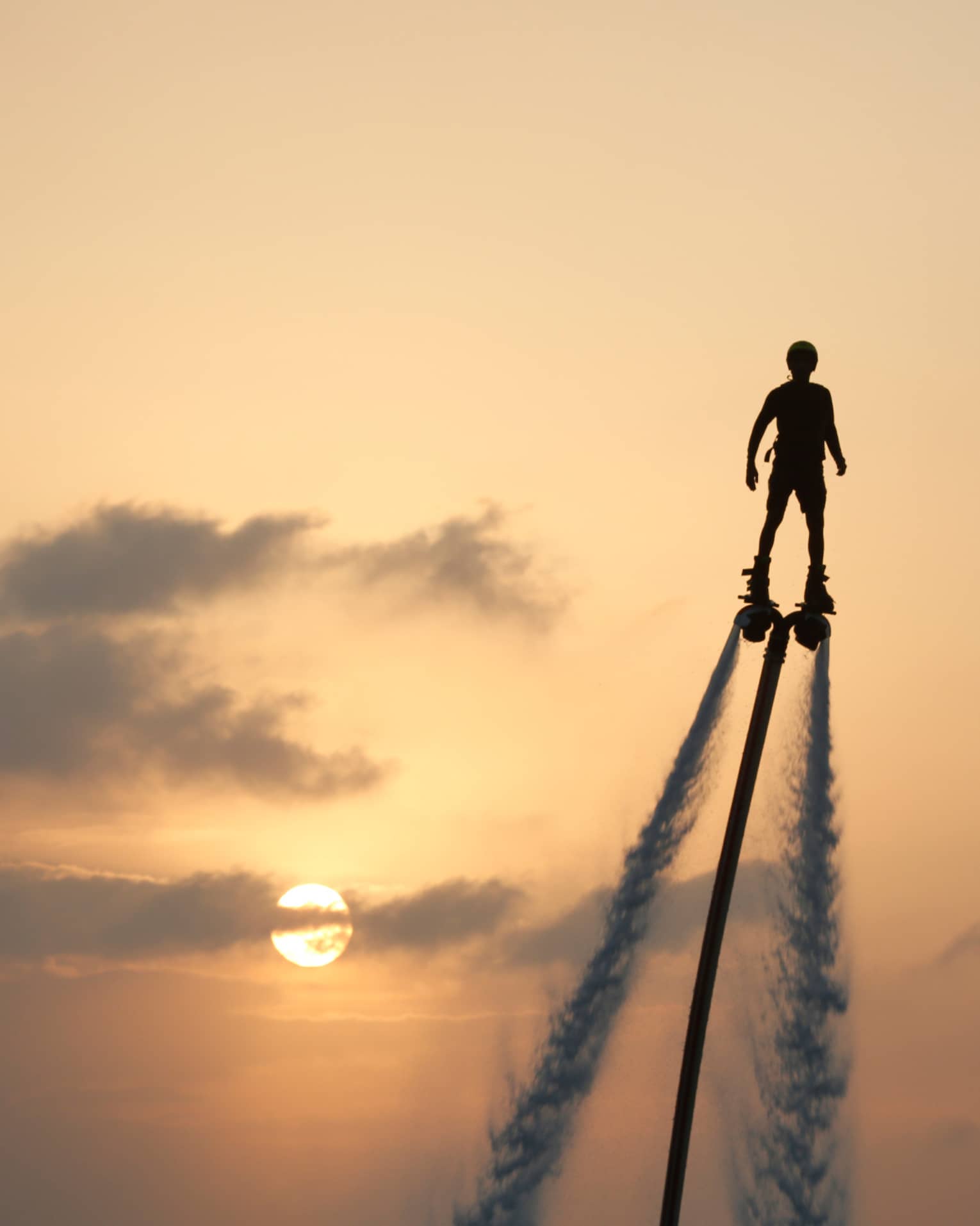 Silhouette of person soaring high above ocean on jet pack, water trails below, against sunset