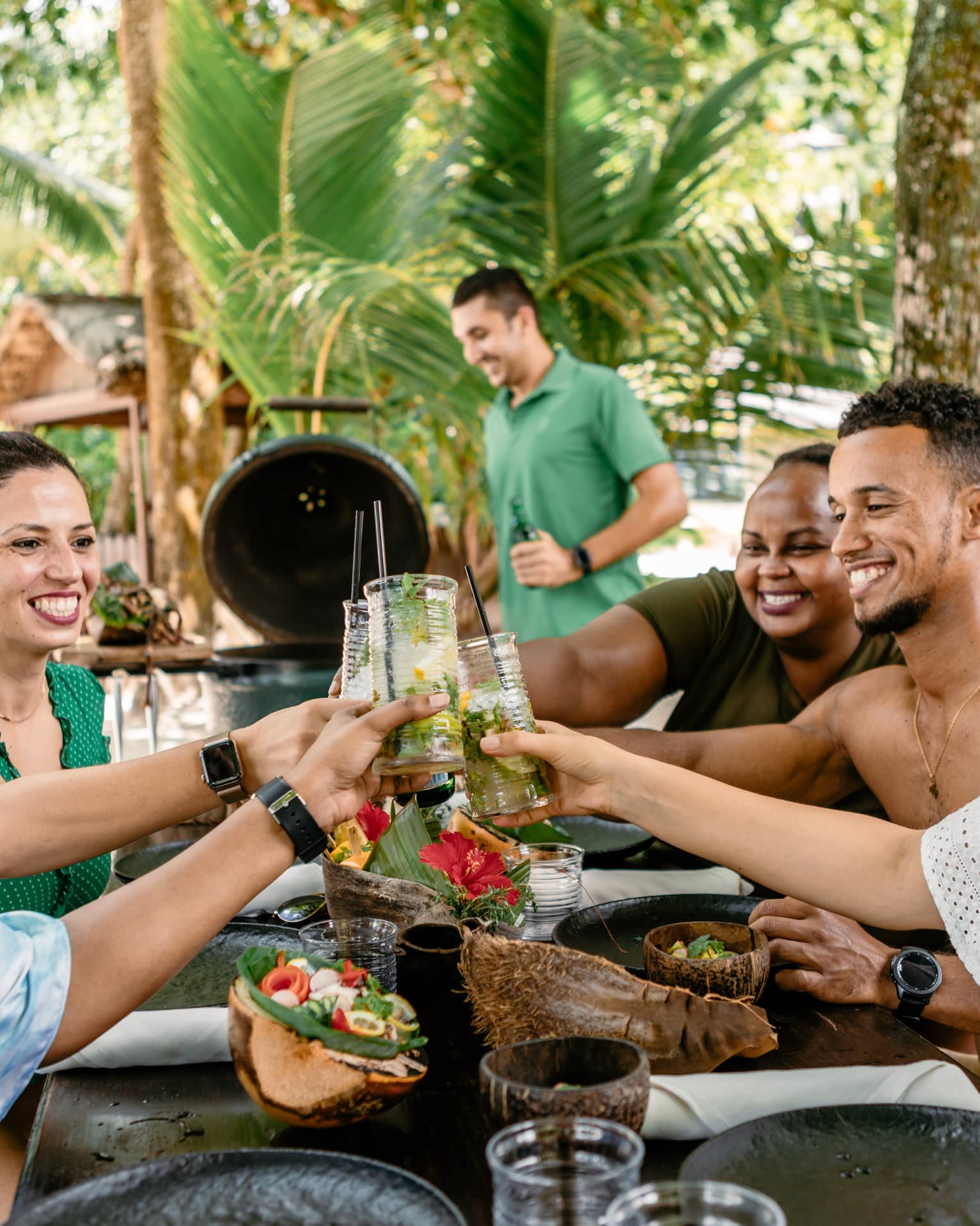 Five friends smiling and clinking glasses over outdoor barbecue lunch in tropical setting