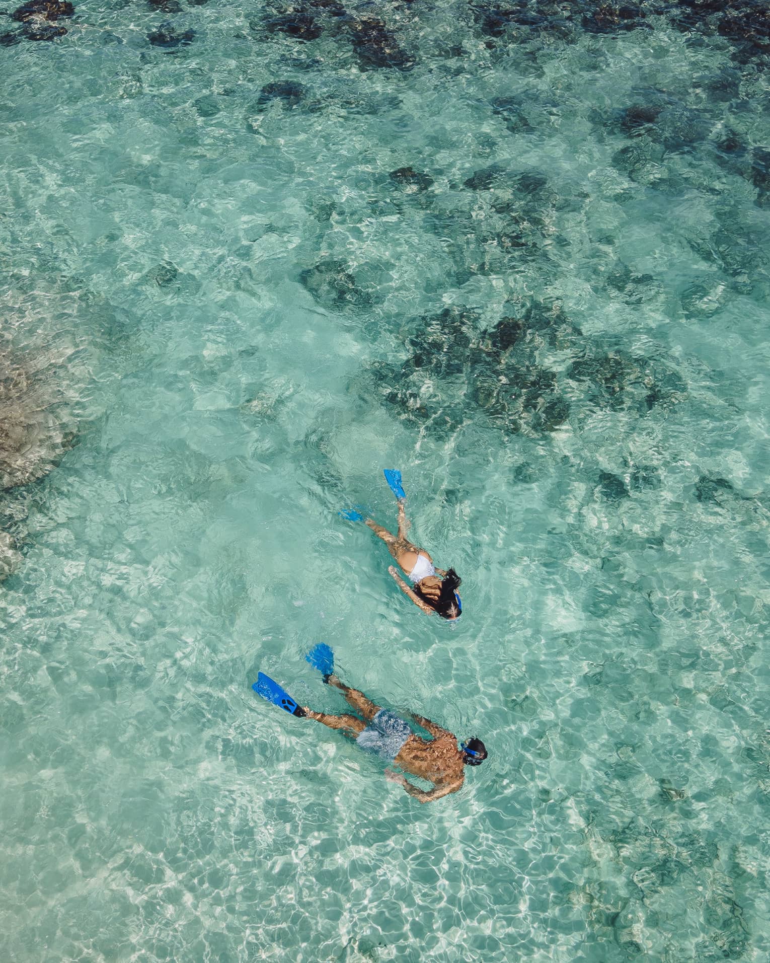 Aerial view of two snorkellers wearing blue flippers in an expanse of shallow, turquoise waters rippling over the reef.