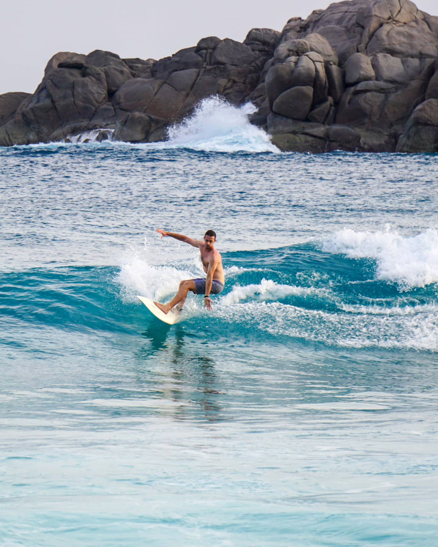 Man on surfboard rides on small blue wave, large rocks in background