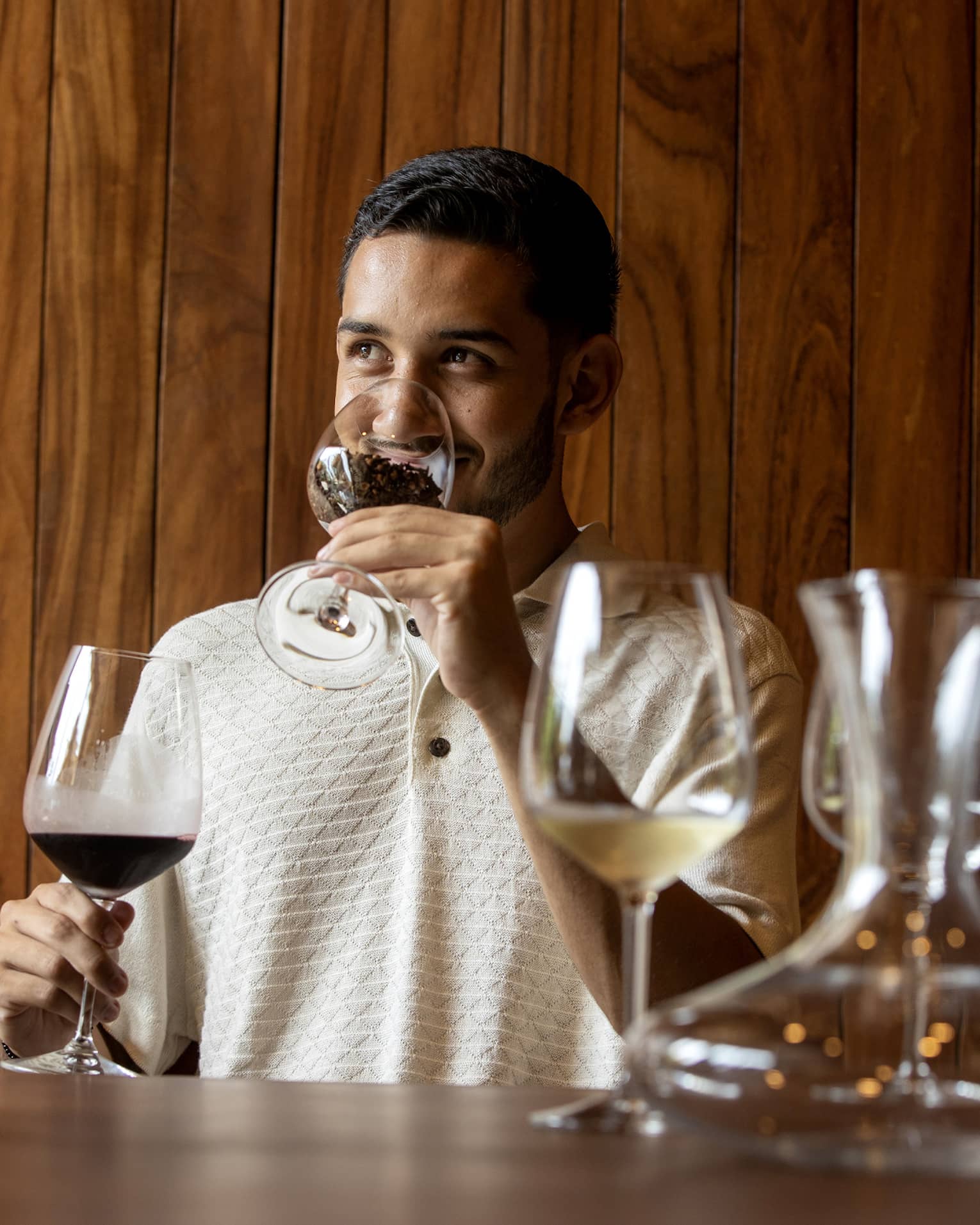 At a wine tasting, a guest smells coffee beans in a glass before tasting the glass of dark red wine held in his other hand. 