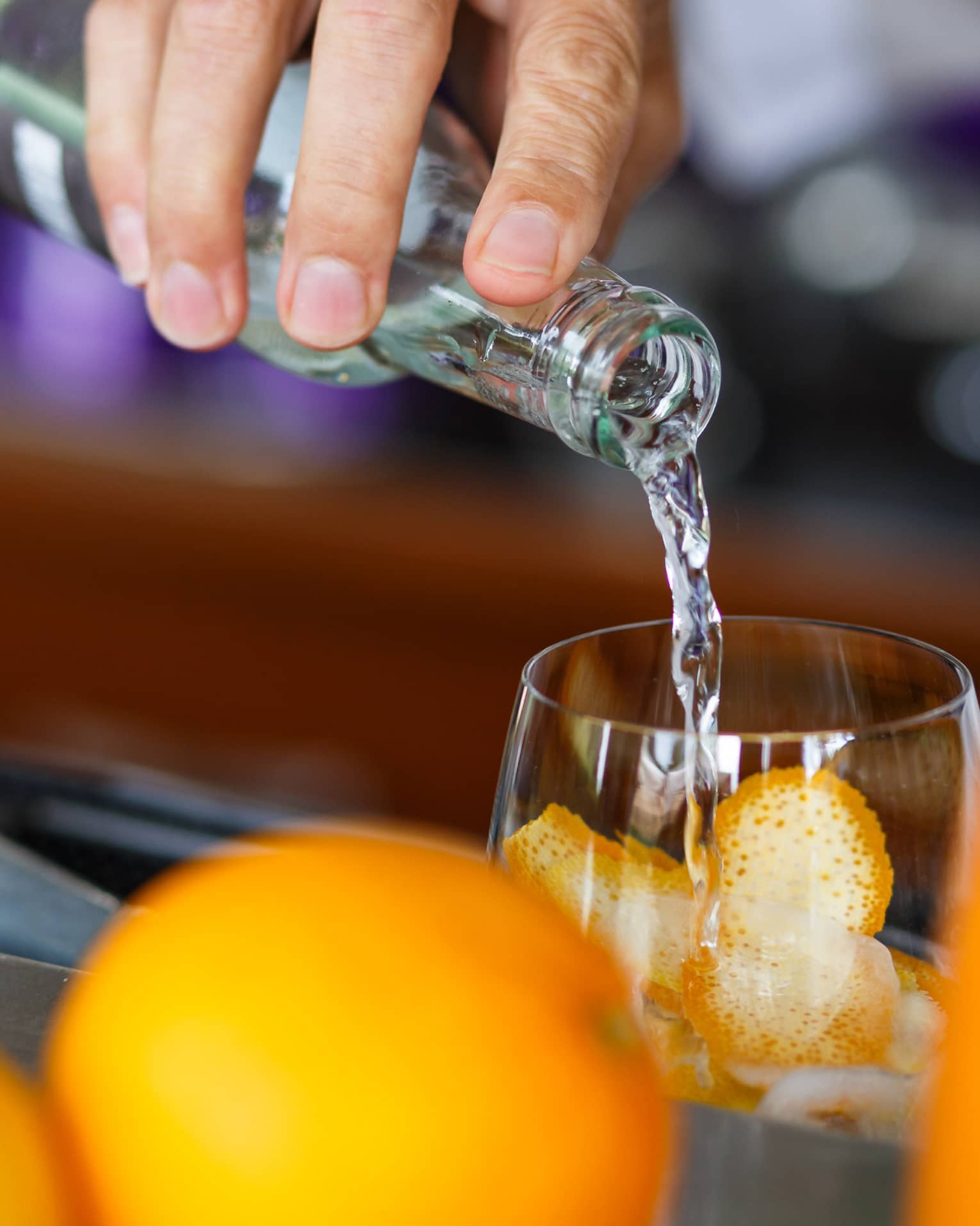 A hand pours clear liquid from a bottle into a glass filled with orange rind; in the foreground, whole oranges and a carafe.