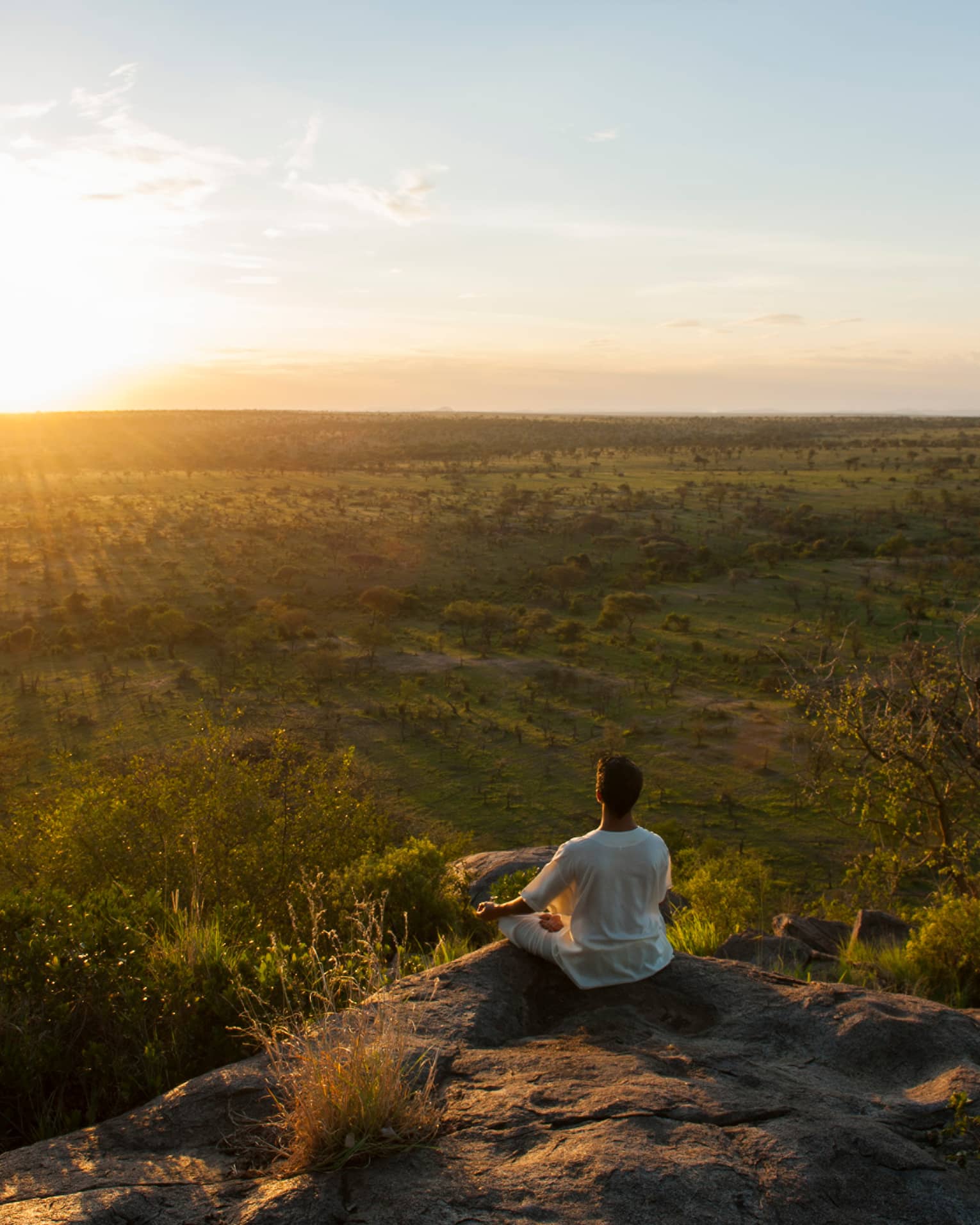 Woman sits crossed-legged, meditates on cliff overlooking Serengeti sunset