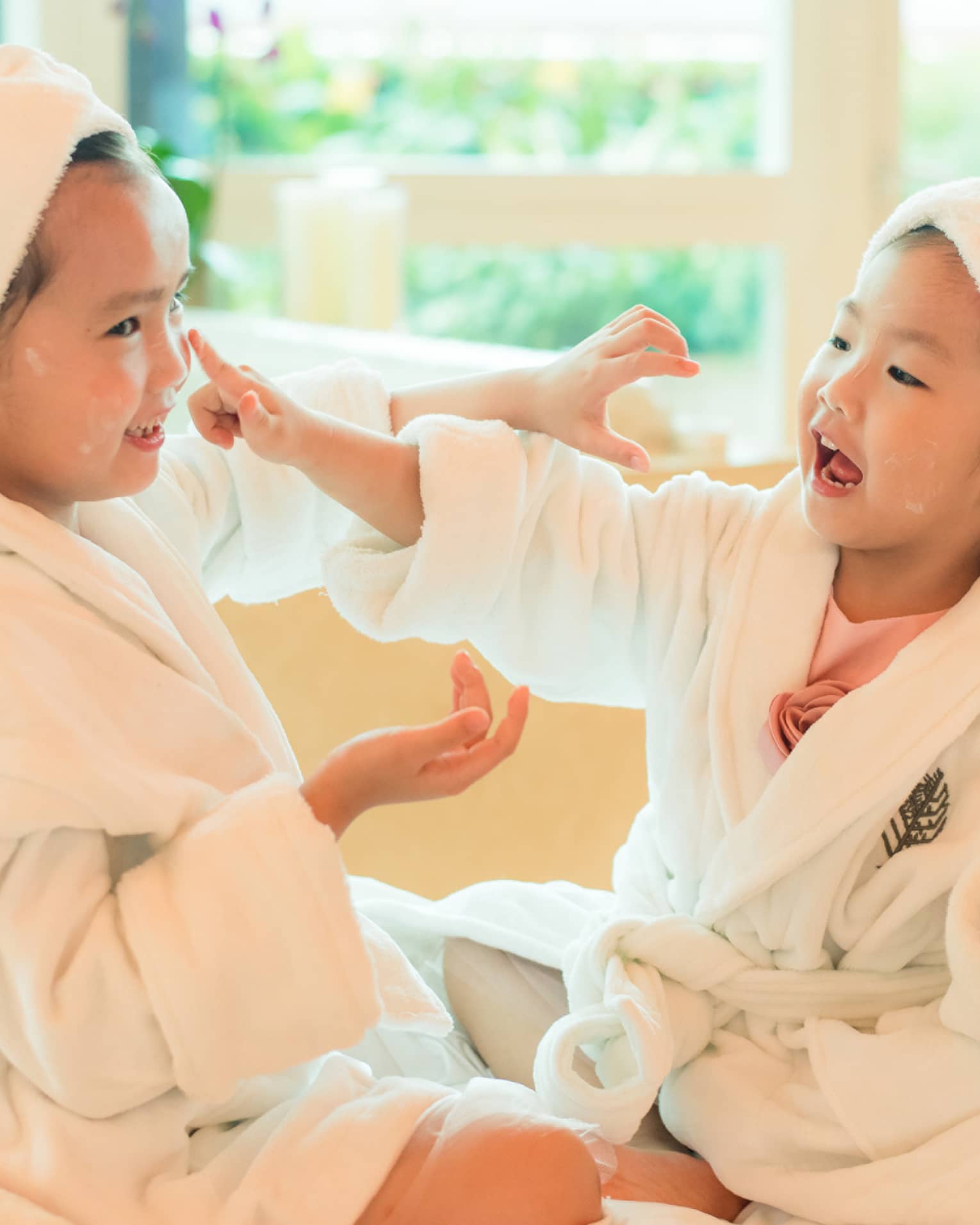 Two laughing young children in white bathrobes with towels around hair put lotion on each other's faces 