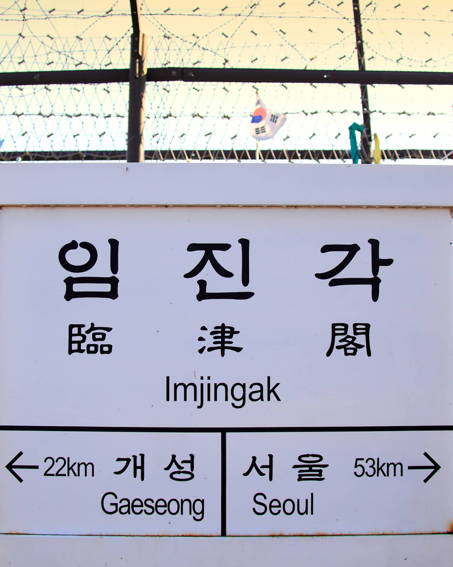 Korean and English text on white sign in front of barbwire fence with ribbons, marking border between North and South Korea.