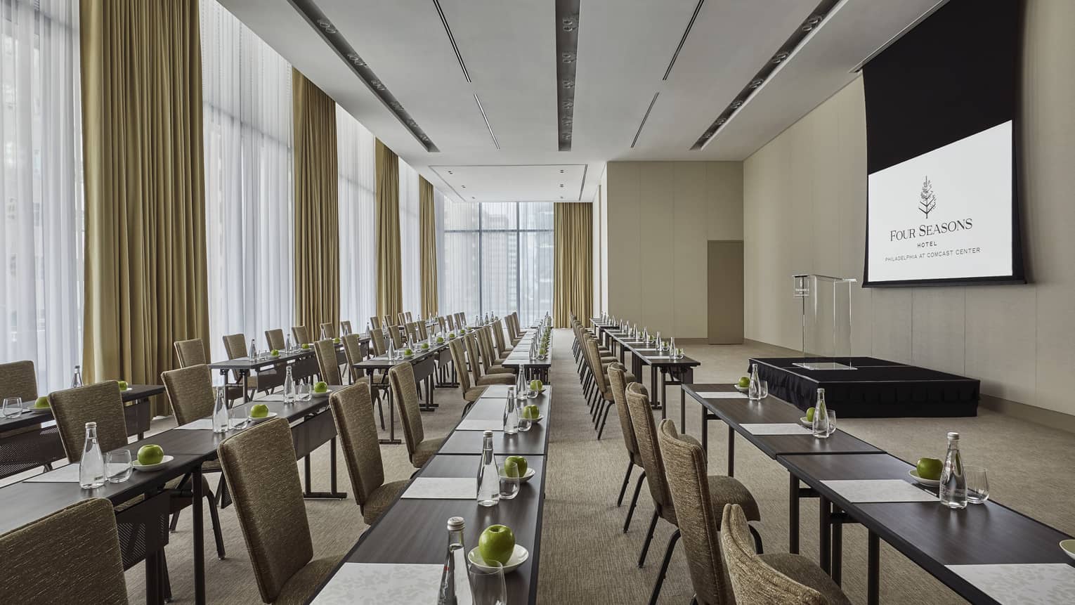 A large presentation room with rows of tables and chairs facing a podium and project screen.