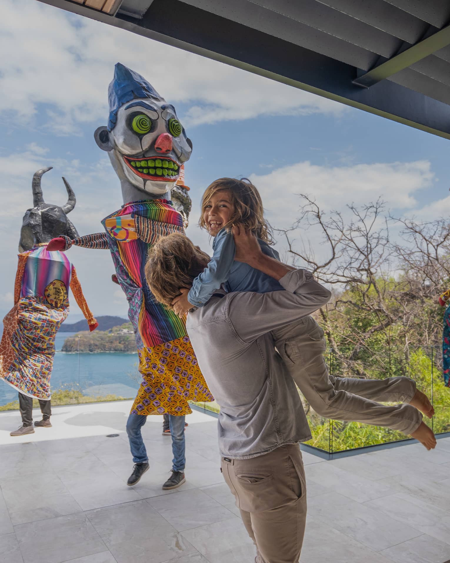 A young boy with blond curly hair is lifted up into the arms of his father as three people dressed in traditional Mexican costumes dance in the background