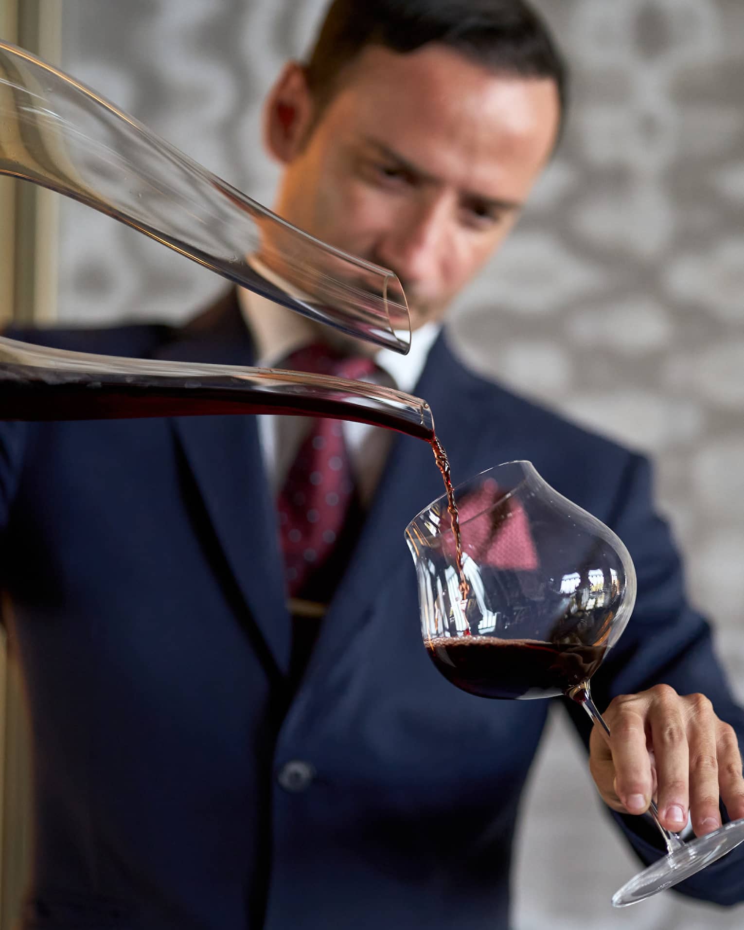 Male in dark blue suit pours a glass of red wine from a large decanter