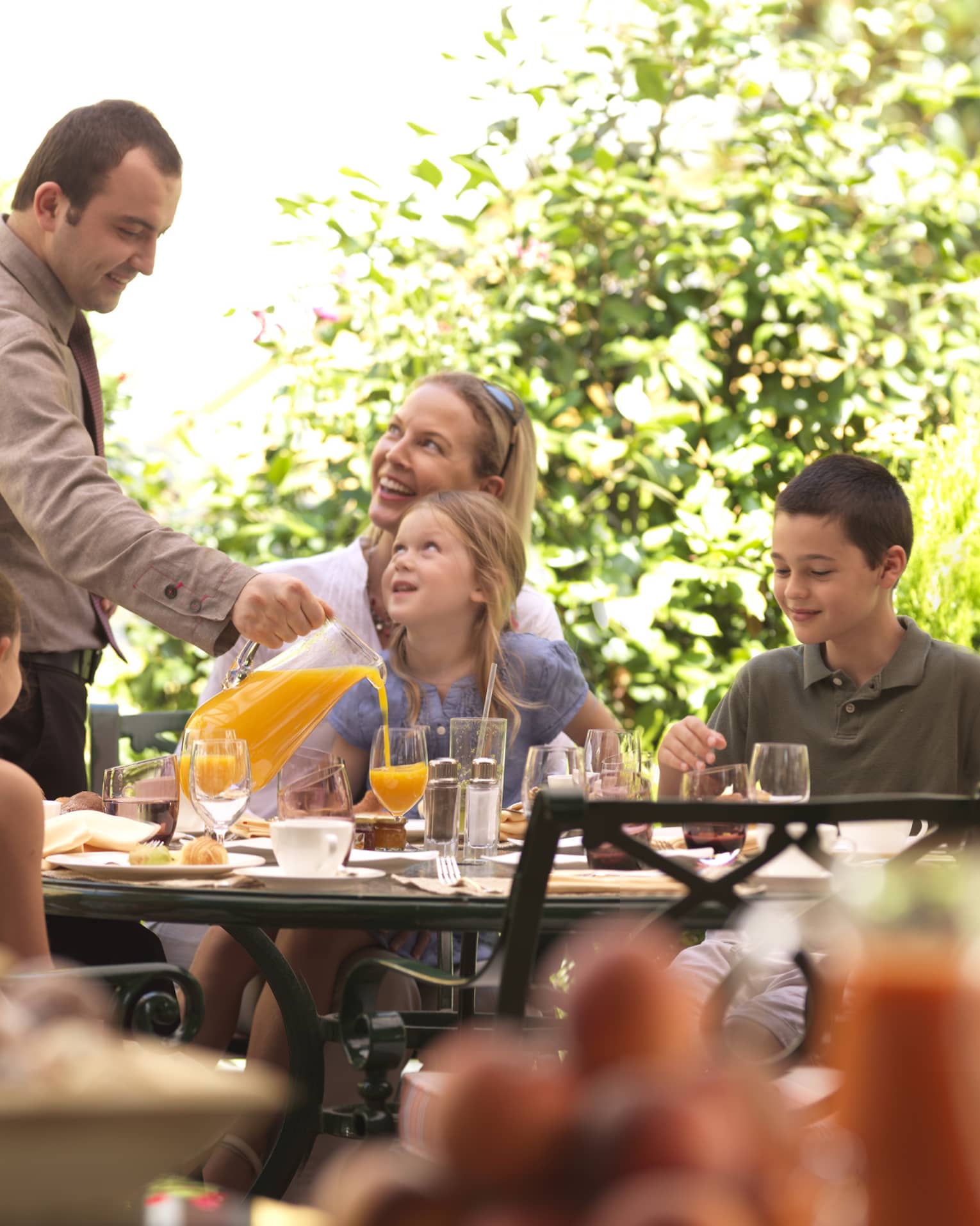 Family brunch on outdoor patio table, staff pours orange juice for young girl 