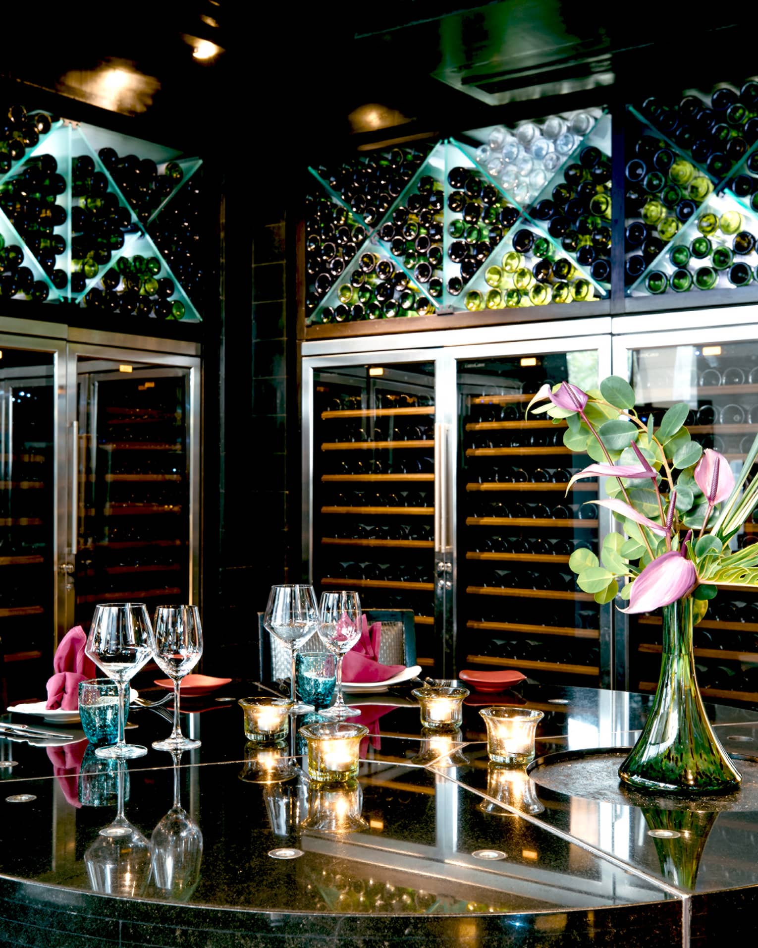 Opulent setting of goblets, candles and a tropical flower atop a mirrored table, surrounded by floor-to-ceiling wine racks.