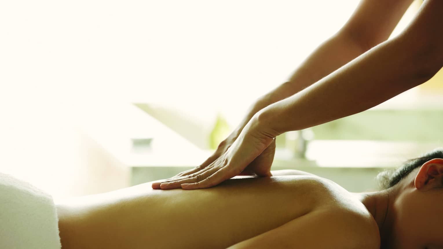 Masseuse reaches over, massages woman's bare back as she lies on spa table