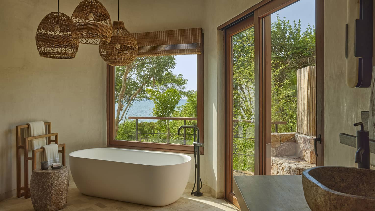 Bathroom with freestanding tub, three hanging whicker lights and window to tropical forest