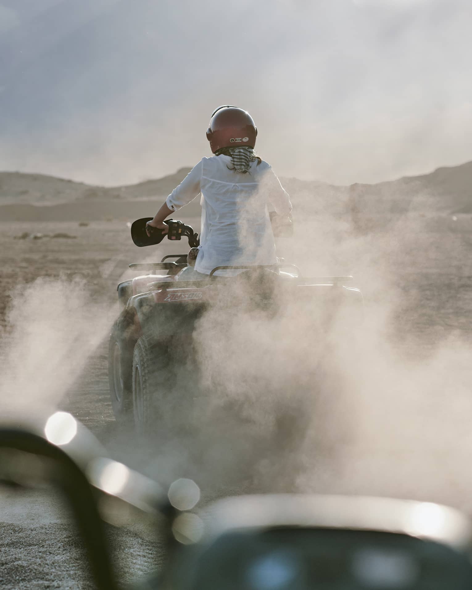 Rear view of an adventurer donning a red helmet while riding an ATV amid dust clouds; desert mountains ahead in the distance.