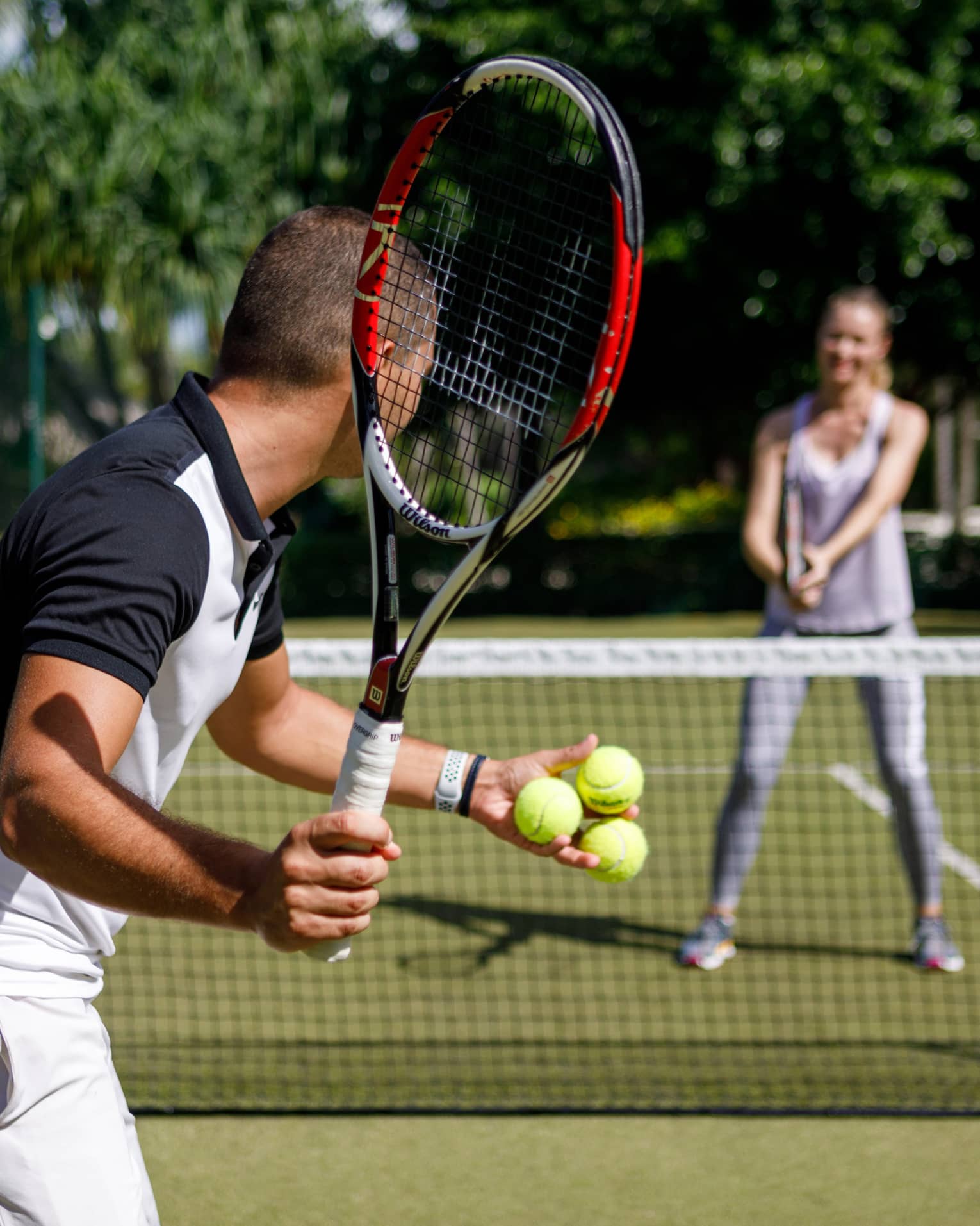 Couple playing tennis on court, man prepares to serve tennis balls to woman ready to swing