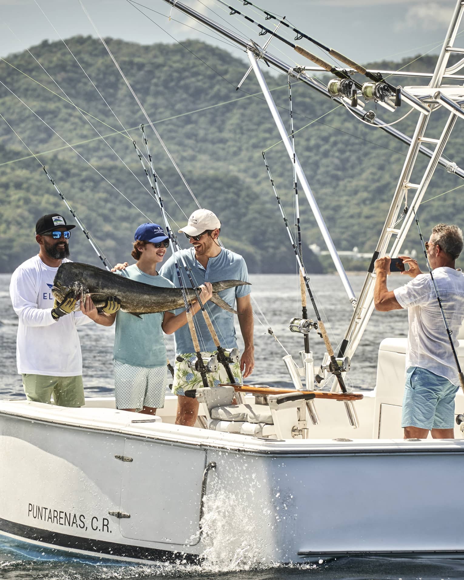 Standing at the back of a fishing boat in the glistening sea against a lush mountain, three smiling people hold a huge fish.