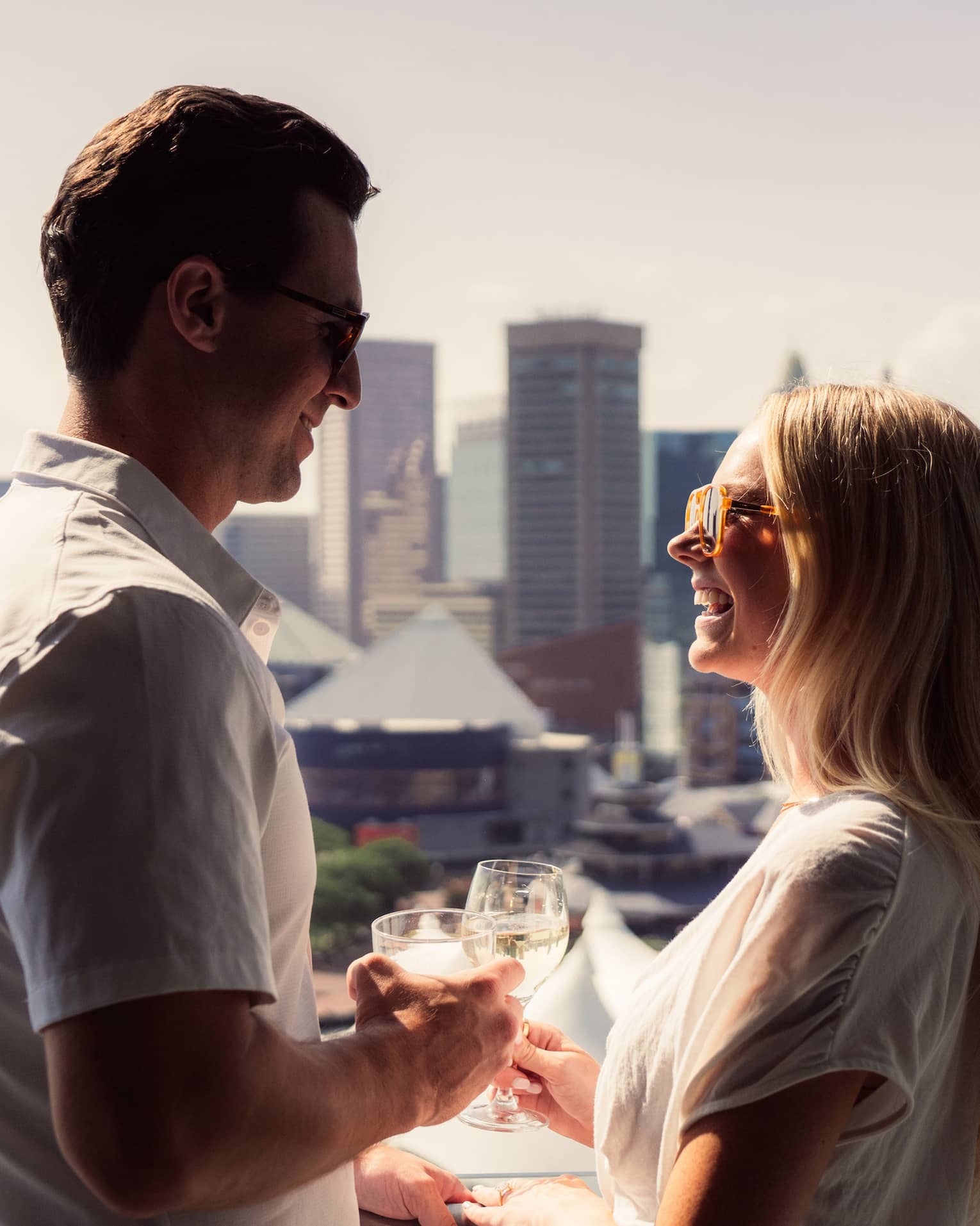 A man and woman drinking on a balcony overlooking water and a city.