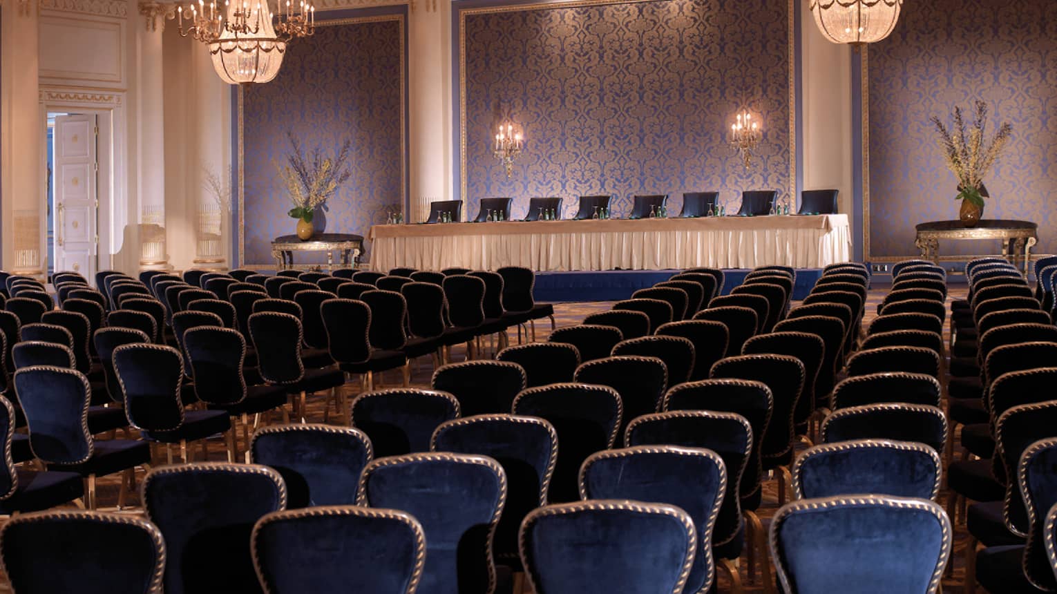Ballroom with royal blue chairs in rows facing podium with table, textured blue panels, chandeliers 