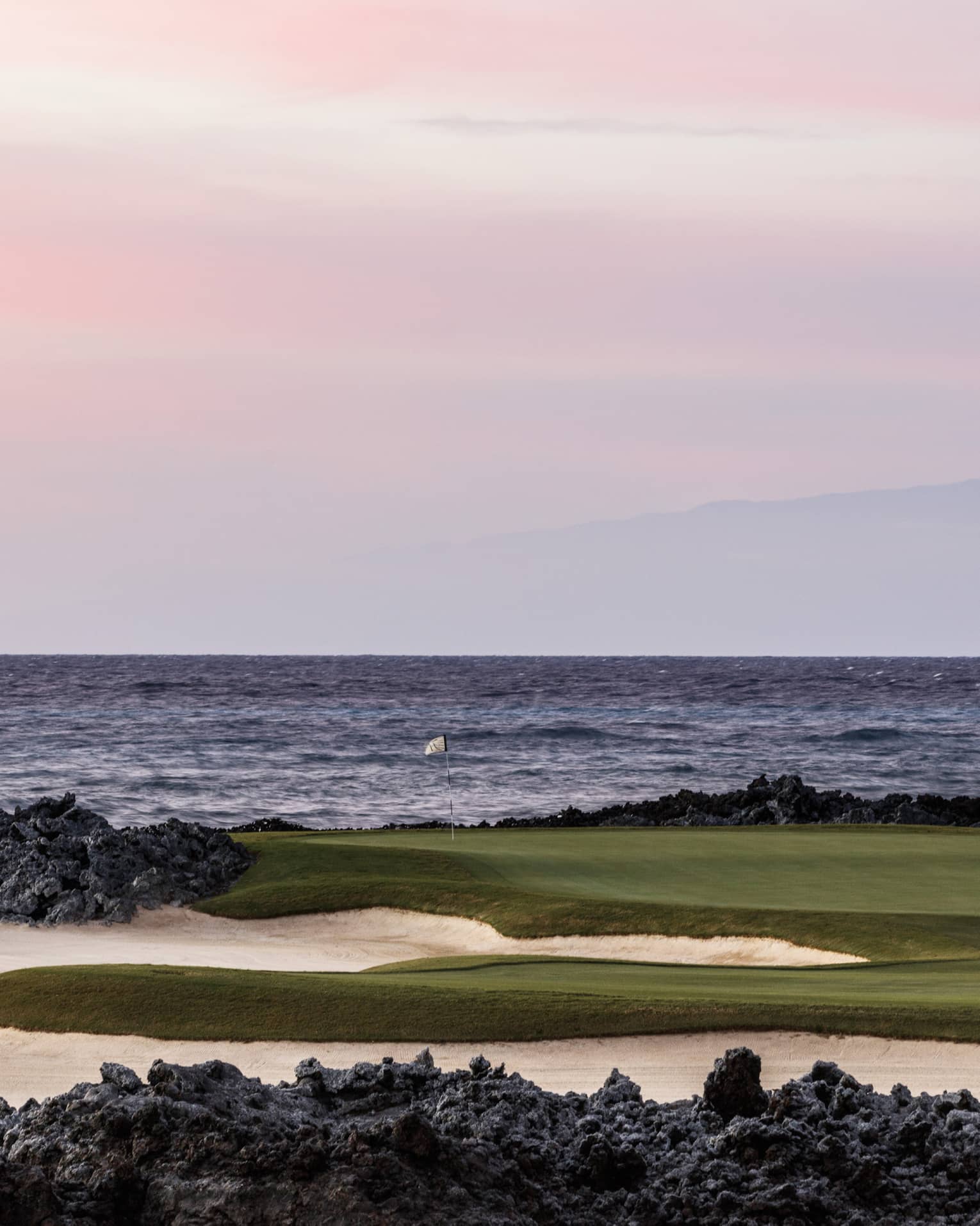 On an oceanside golf course beneath a rose-coloured sky, a single flagstick stands amid black lava rocks and white sand. 