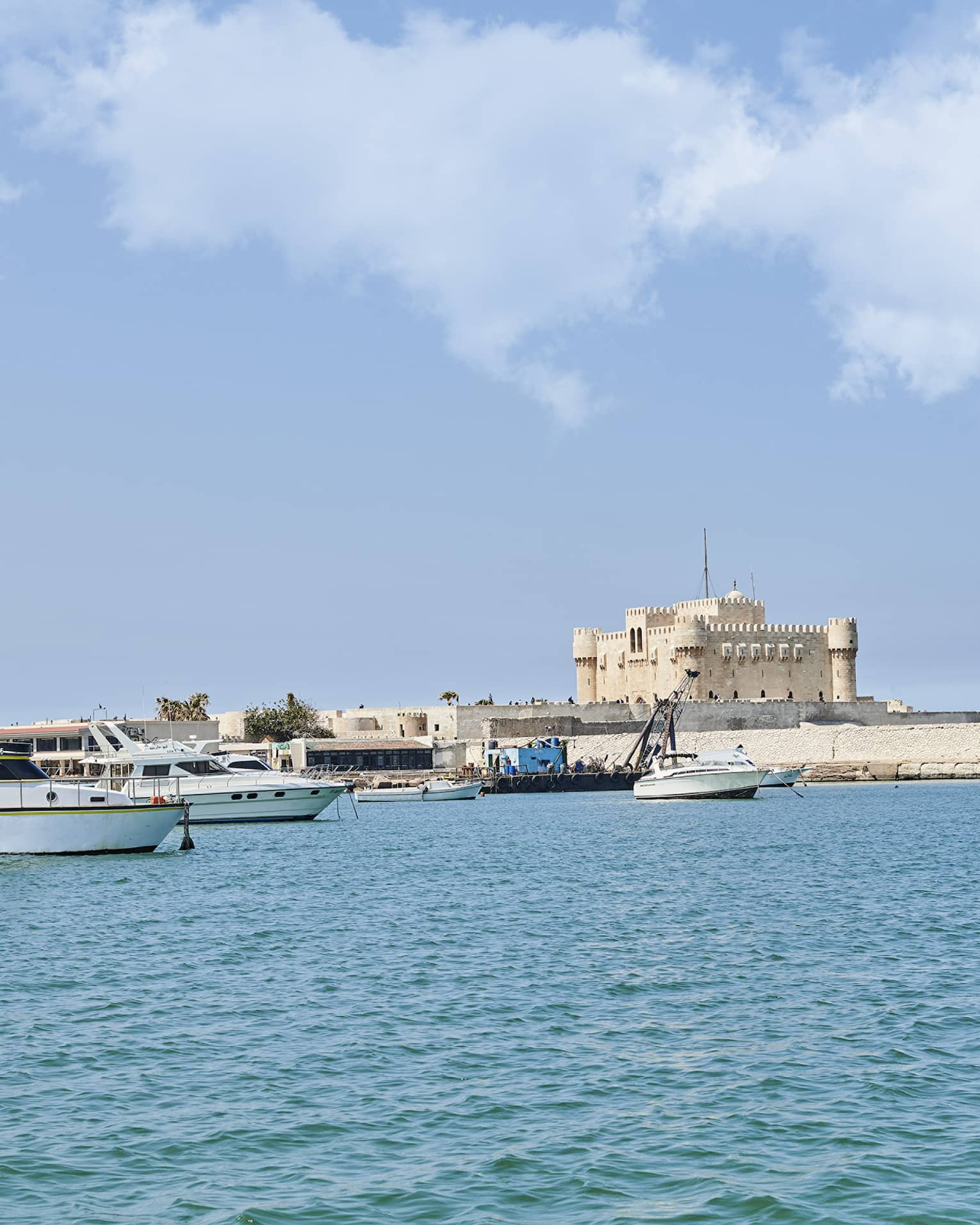 A distant sand-coloured fortress perched on a quay along a shore dotted with small yachts and fishing boats over calm waters.