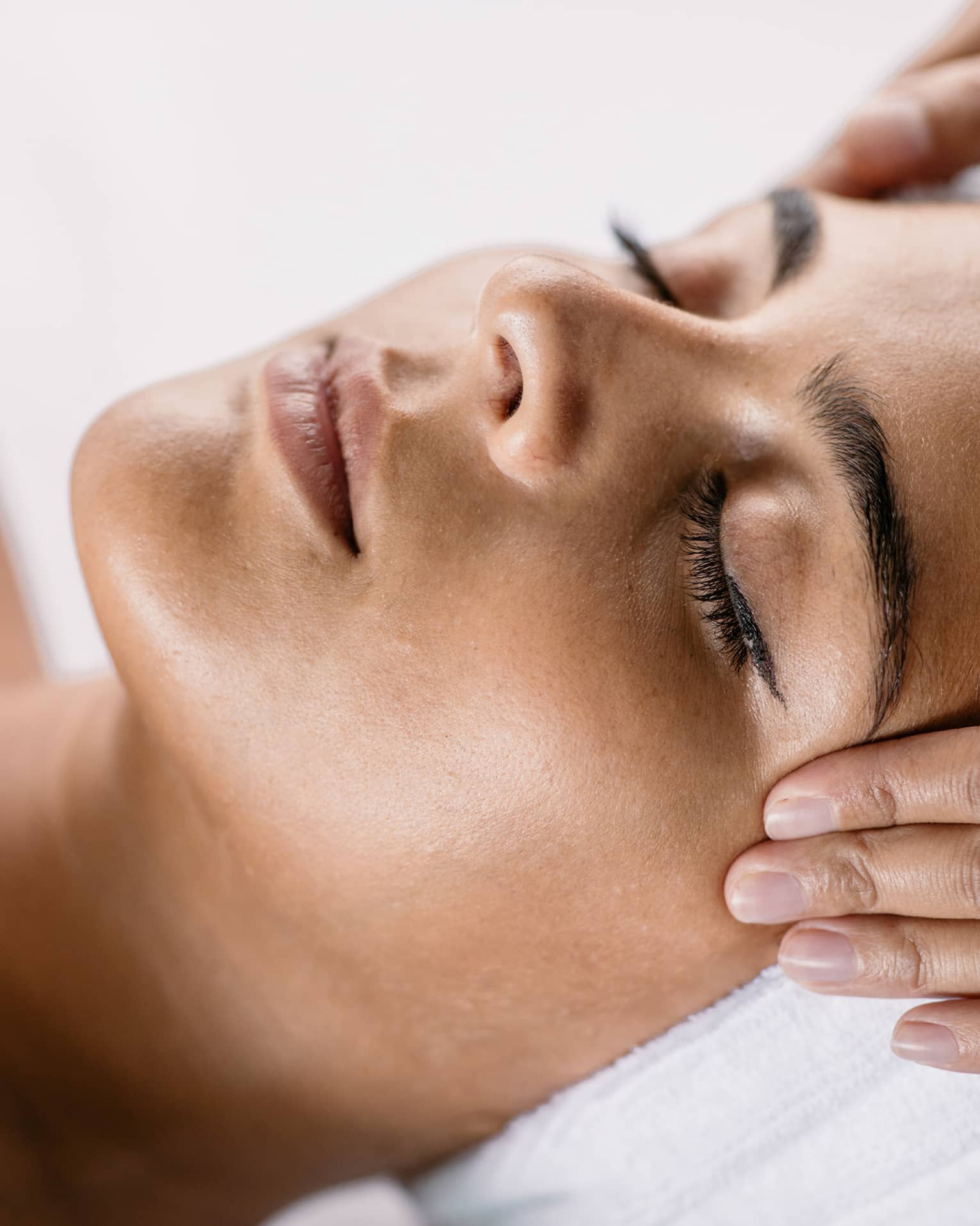 Hands massage woman's temples as she closes her eyes, spa towel around hair
