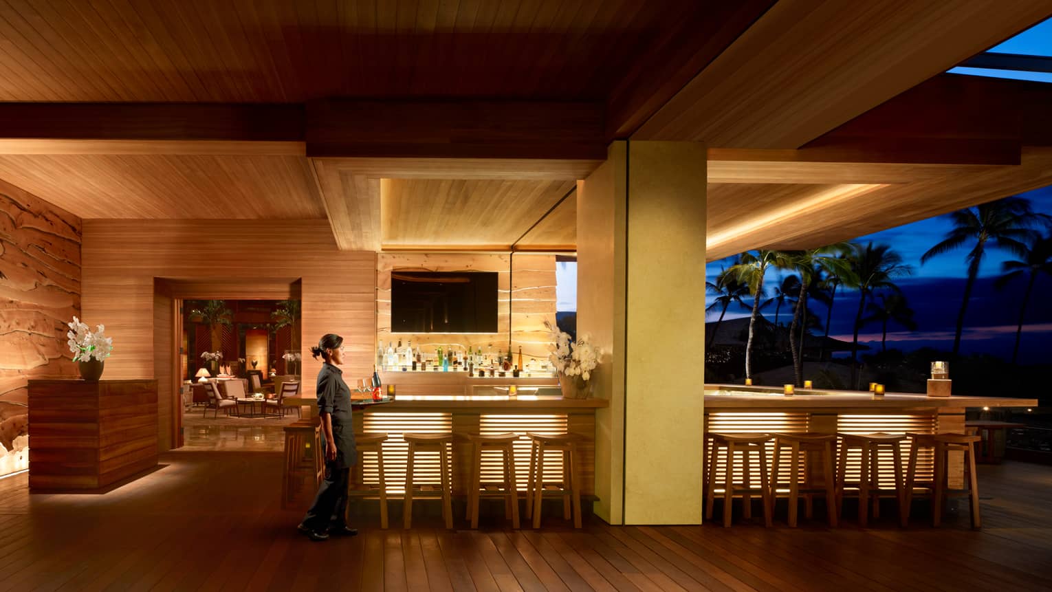 Server walks by wood NOBU Lanai restaurant bar with lights on indoor outdoor patio at night